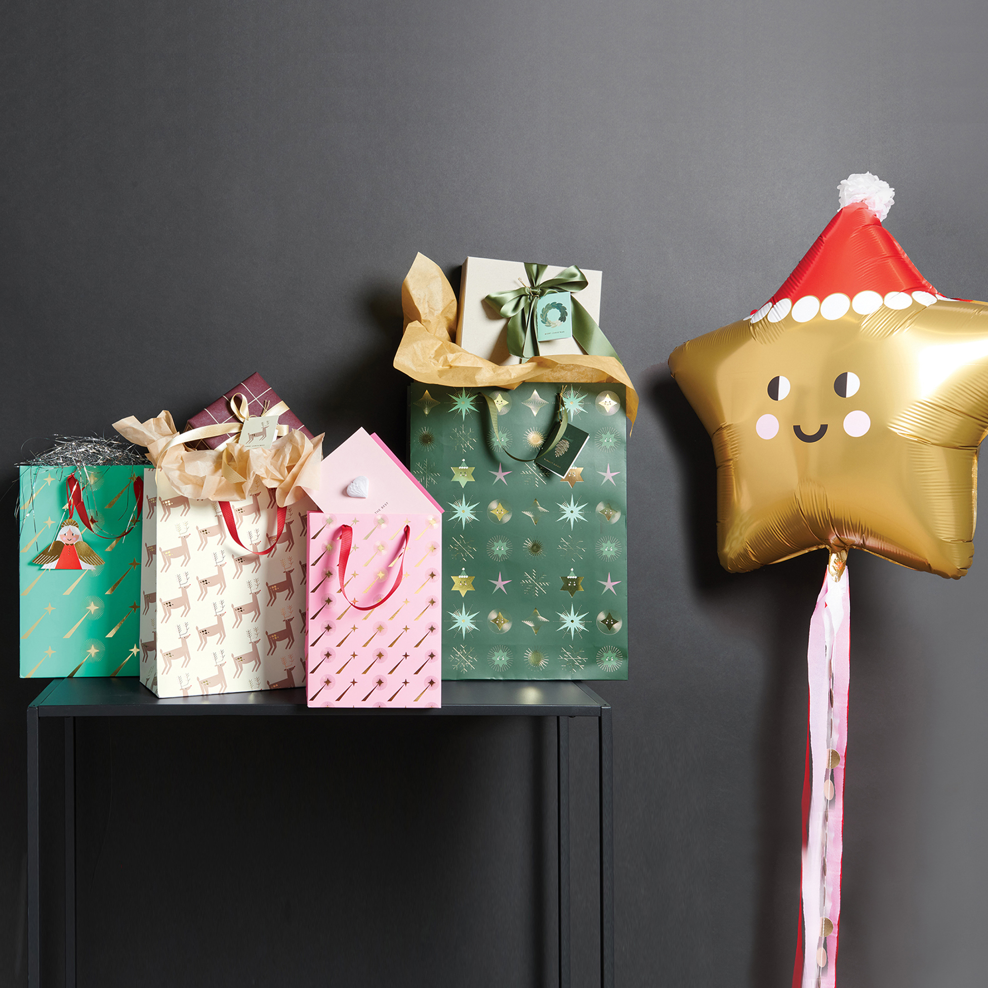 Gefüllte Geschenktüten mit Weihnachtsdesigns auf Tisch vor schwarzer Wand gereiht. Daneben ein Ballon in goldener Sternenform mit Gesicht und Weihnachtsmütze. 