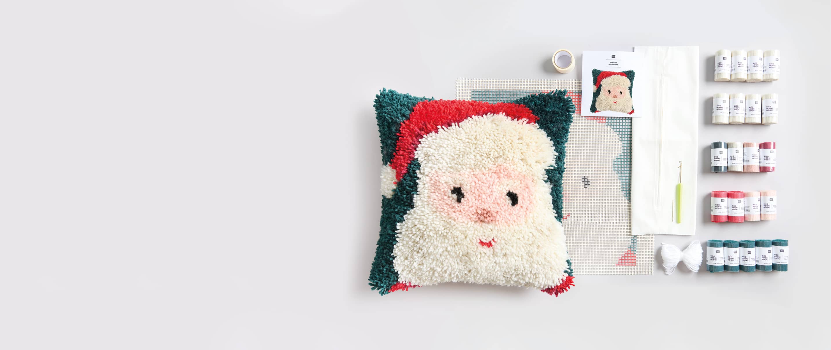 Ein geknüpftes Kissen mit Santamotiv, Knüpfgarn, Haken und weiteres Knüpfzubehör liegen ausgebreitet auf grauem Hintergrund. 