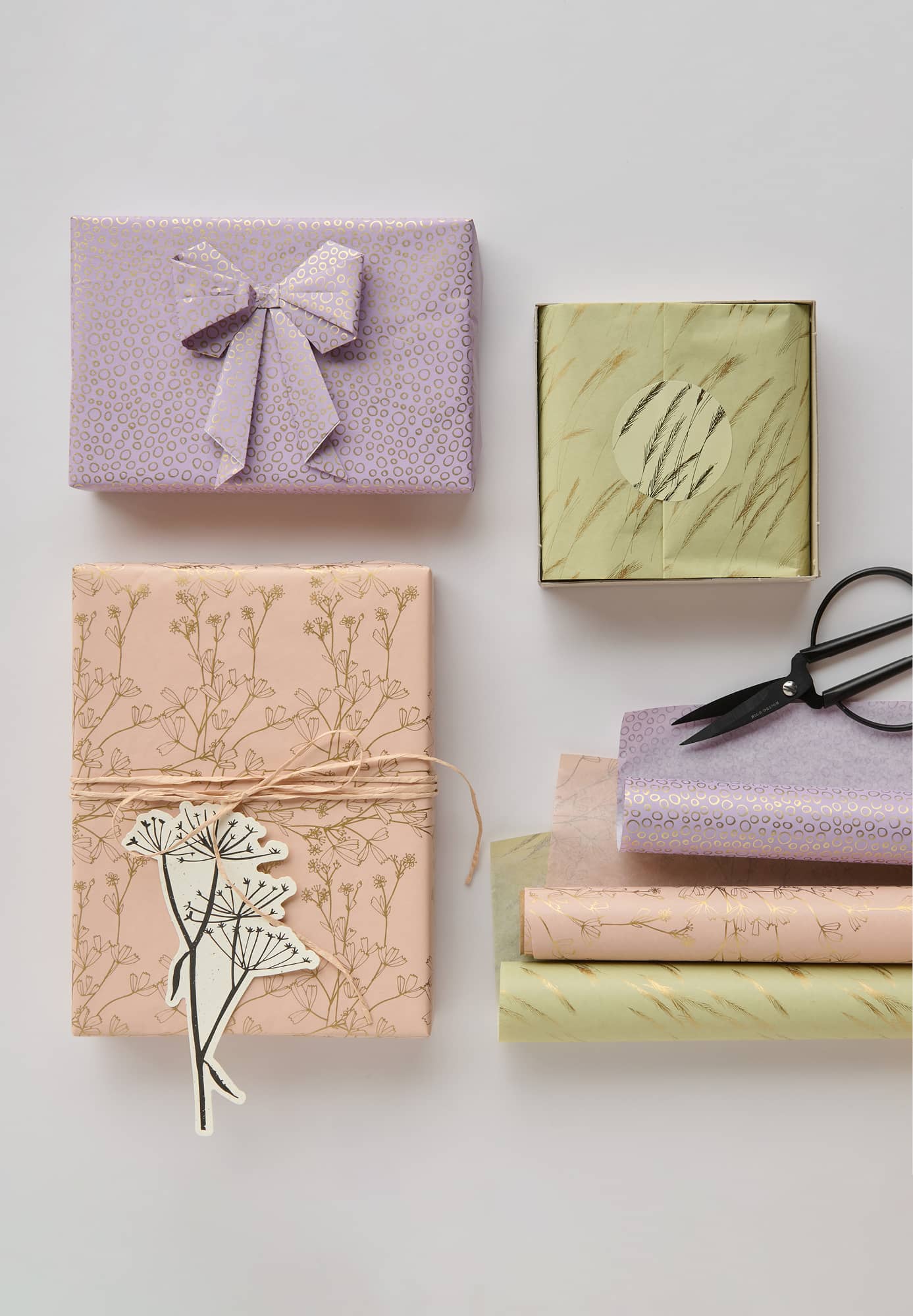 Verpackte Geschenke in den Farben grün, apricot und lila mit goldenen Details liegen zusammen mit Seidenpapierrollen und Schere auf grauem Hintergrund. 