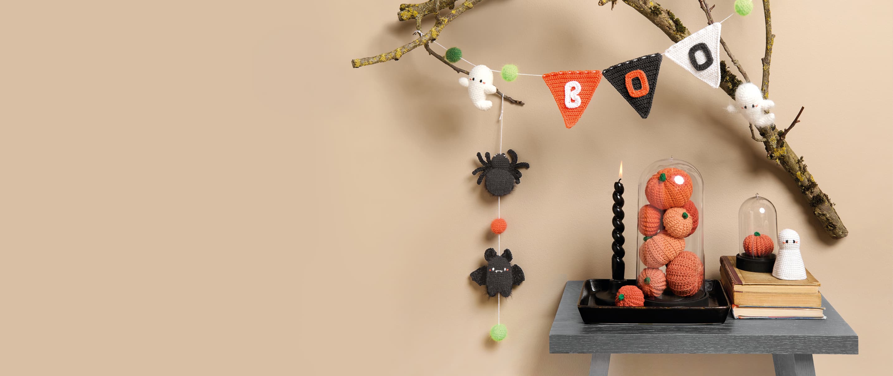Gehäkelte Halloweenfiguren in Form von Kürbissen, Gespenstern und Spinnen sind auf einem Tischchen und an einem Ast dekoriert. 