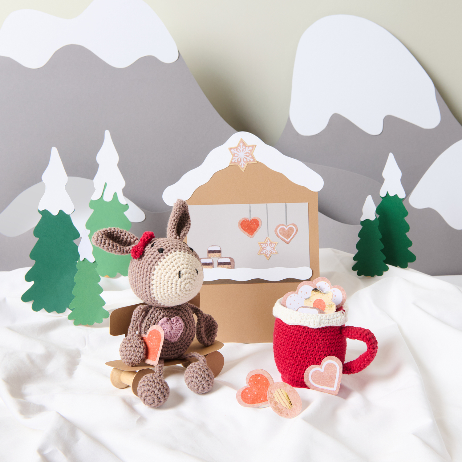 Häkelanleitung Ricorumi CAL Daisy the Christmas Cookie Donkey: Ein gehäkelter Esel sitzt vor einer winterlichen Kulisse mit Tannen und Lebkuchenhaus.