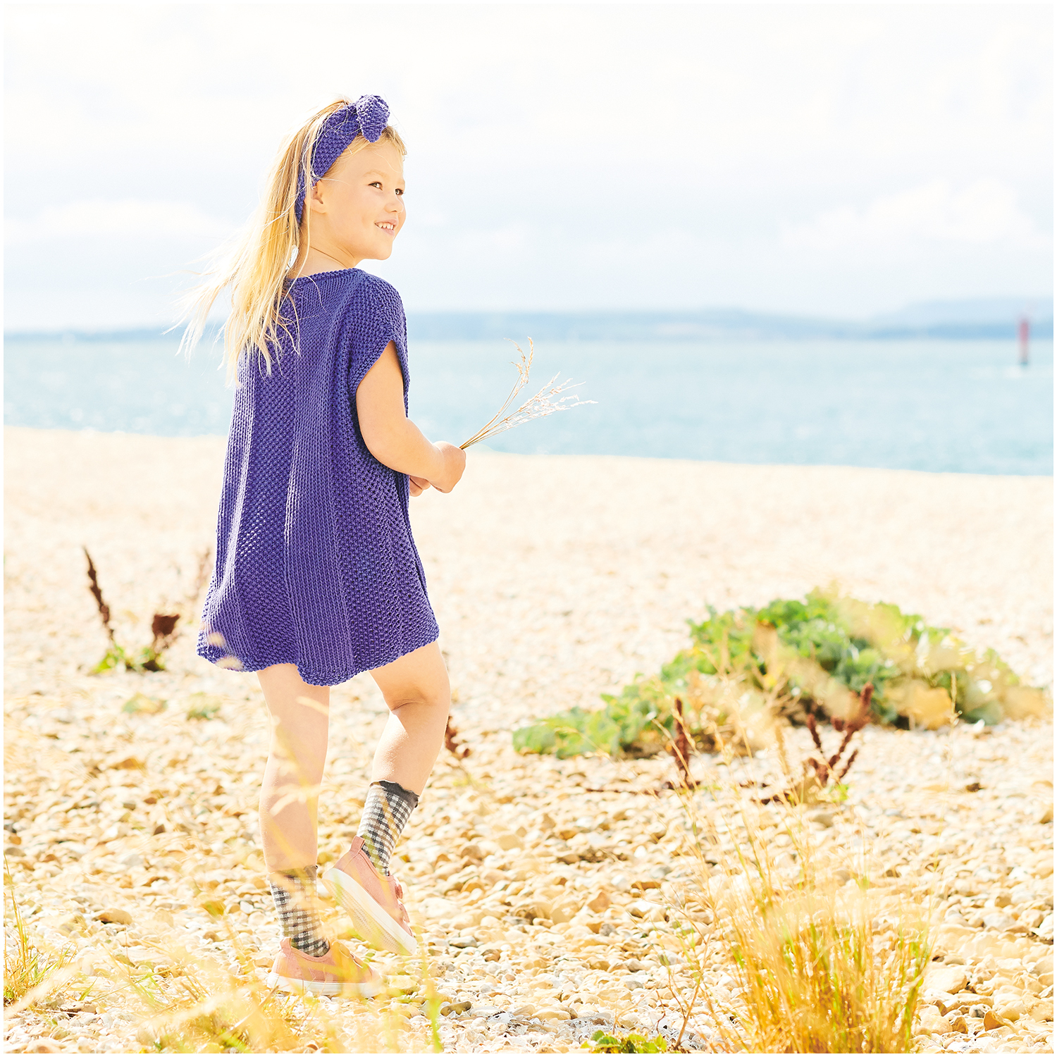 Essentials Organic Cotton aran: Mädchen in violettem Kleid am Strand, lächelnd und mit Blumen in der Hand.