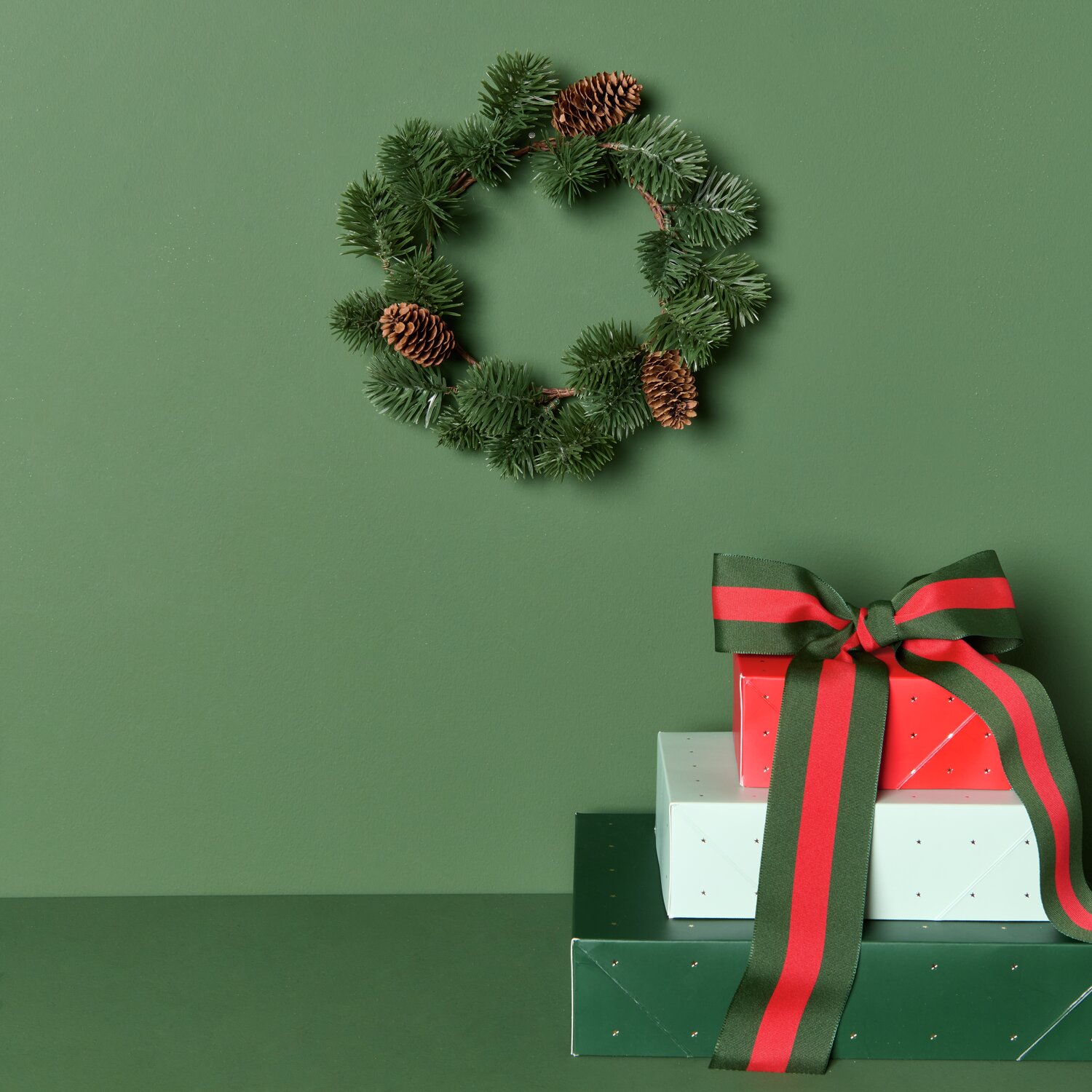 Paper Poetry Papierschachtel Sterne Set Mix, Tannenkranz mit Zapfen auf grünem Hintergrund, daneben Geschenke mit Schleifen.