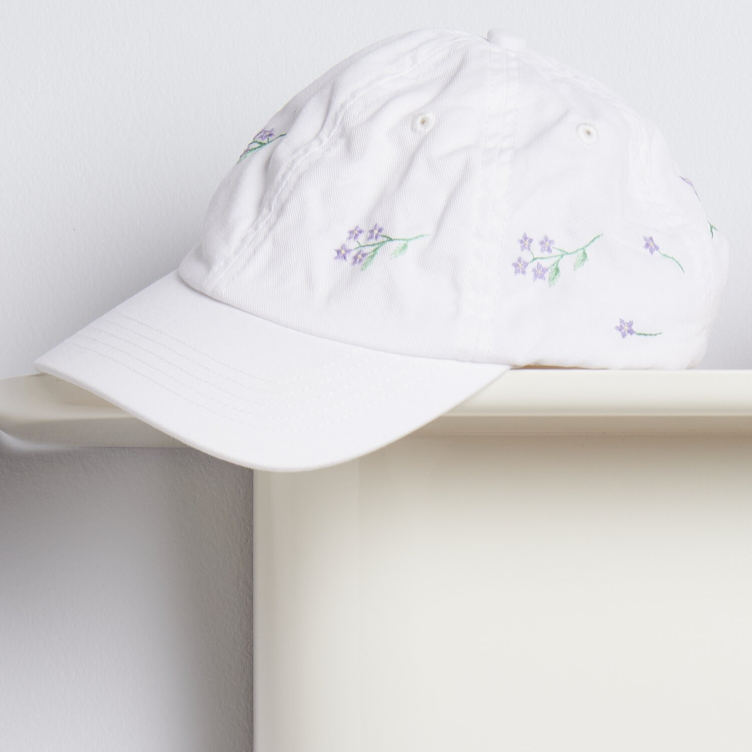 Stick & Stitch Packung Blumenkränze XL 13 Stück: Weiße Kappe mit lila Blumenstickerei auf einem Regal.