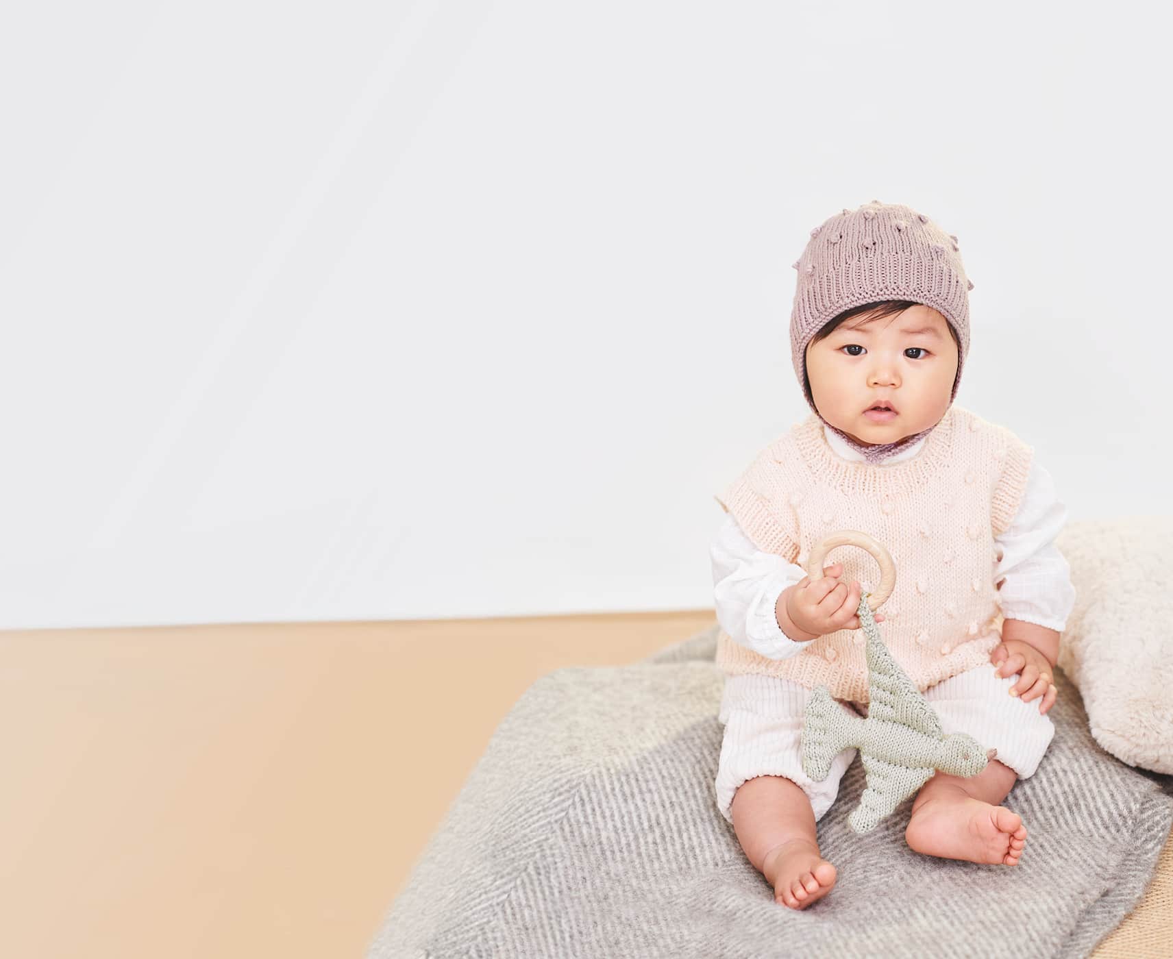 Ein Baby sitzt mit gestricktem Pullover und gestrickter Mütze auf einer Decke und hält einen Beißring in der Hand