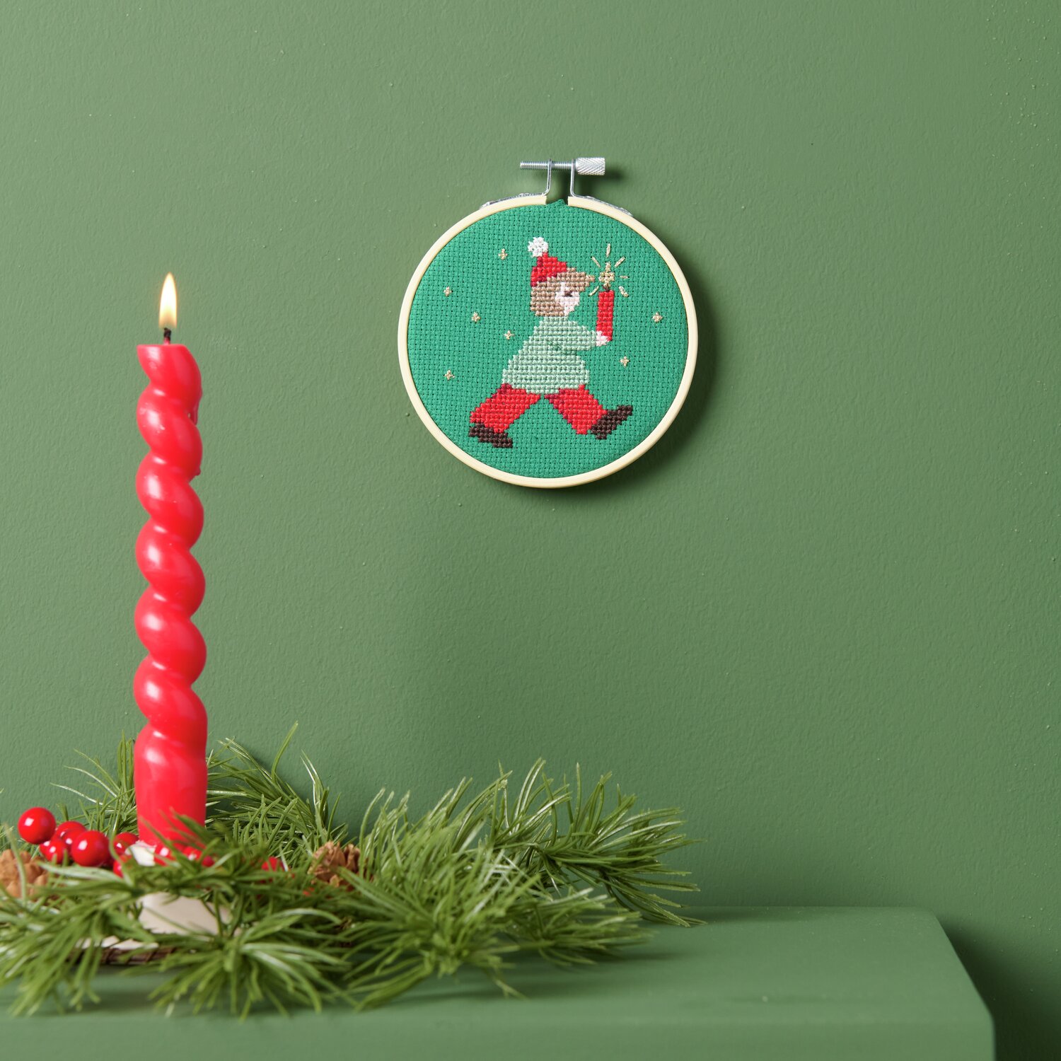 Stickpackung Lichterkind Ø10cm von Rico Design Stickpackung Lichterkind: Ein grüner Stickrahmen mit weihnachtlichem Motiv neben einer roten Kerze auf grünem Hintergrund.