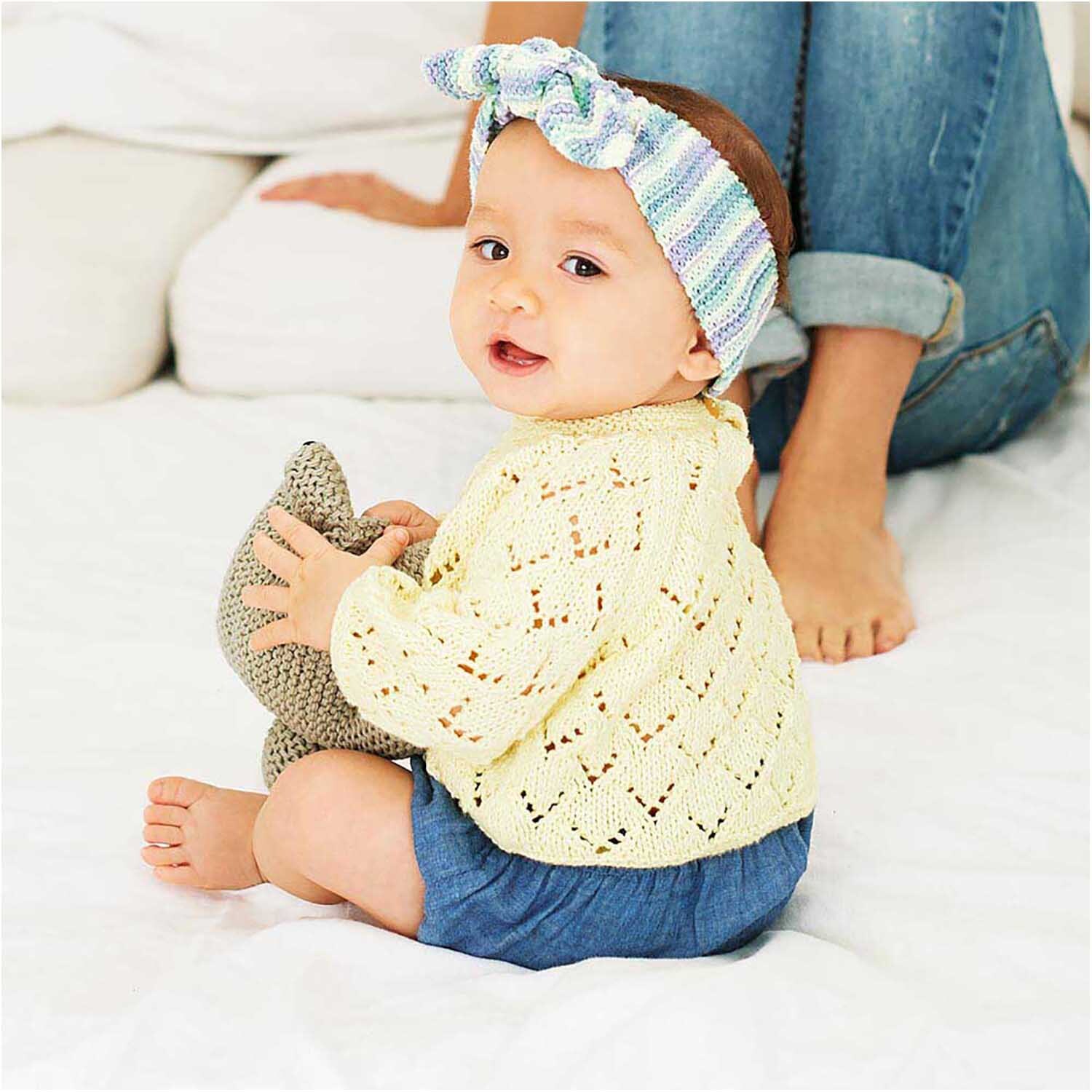 Rico Baby Cotton Soft dk: Ein Baby in pastellgelbem Strickpullover spielt auf einer weißen Decke.