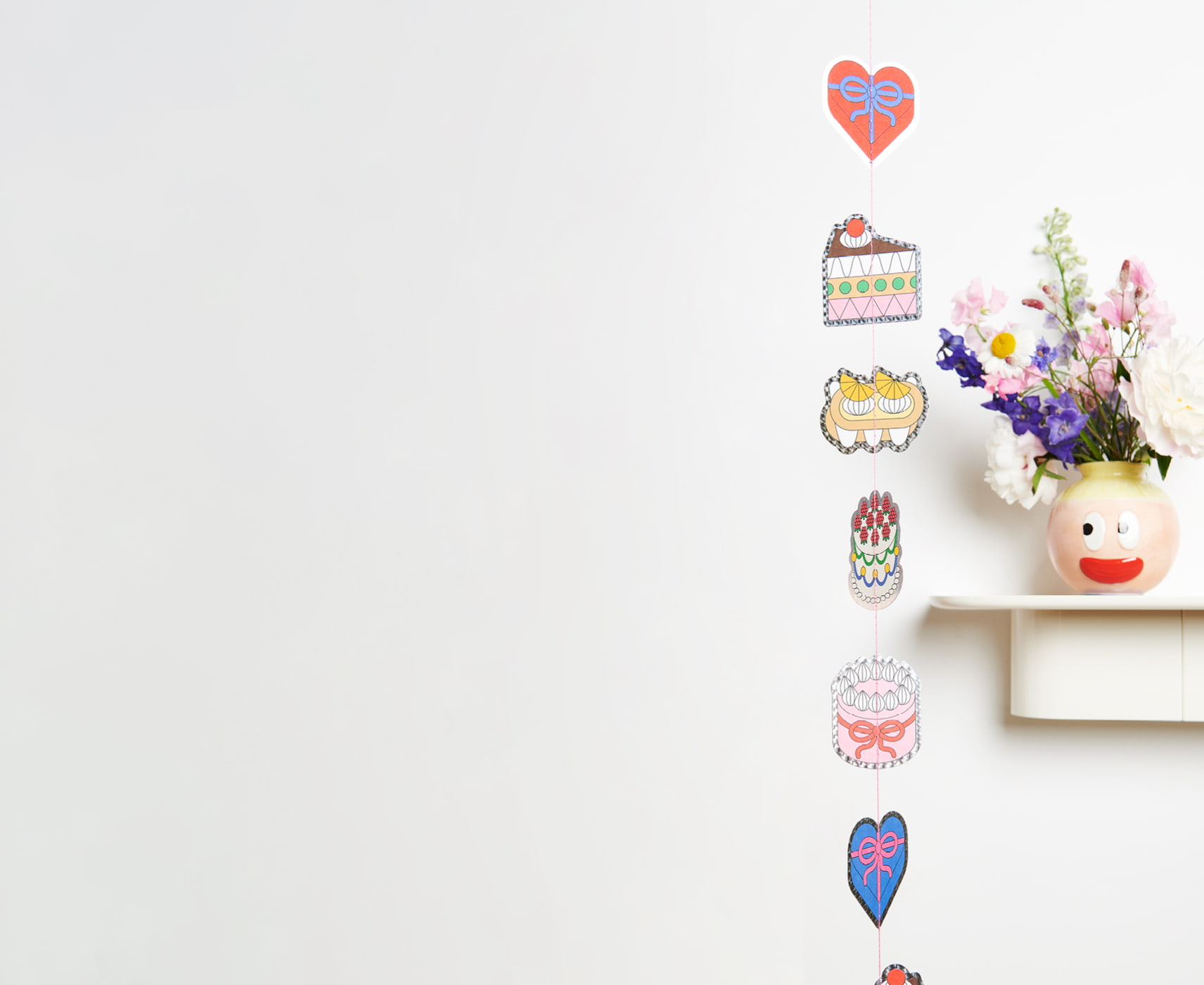 Eine Girlande mit Motiven von Torten und Kuchen hängt an einer Wand neben einer dekorierten Blumenvase.