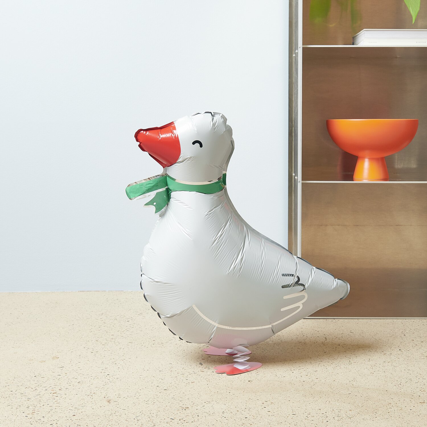 Folienballon Airwalker Gans in Weiß mit grünem Schal, steht auf einem Tisch vor Regal.