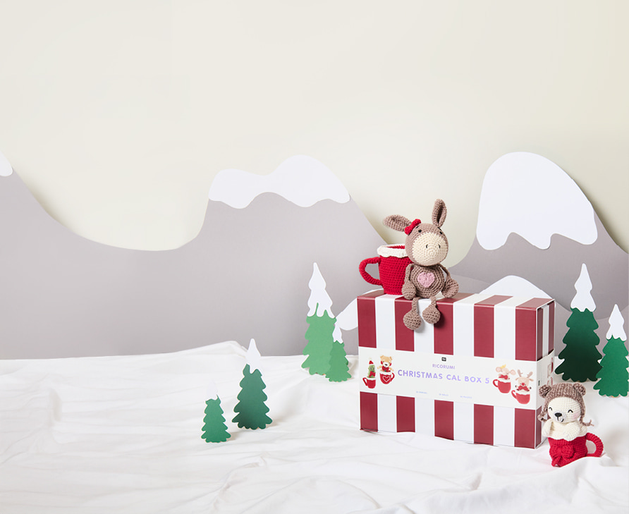 Ricorumi Xmas CAL Box 5 steht in einer Winterlandschaft umgeben von zwei gehäkelten Figuren 