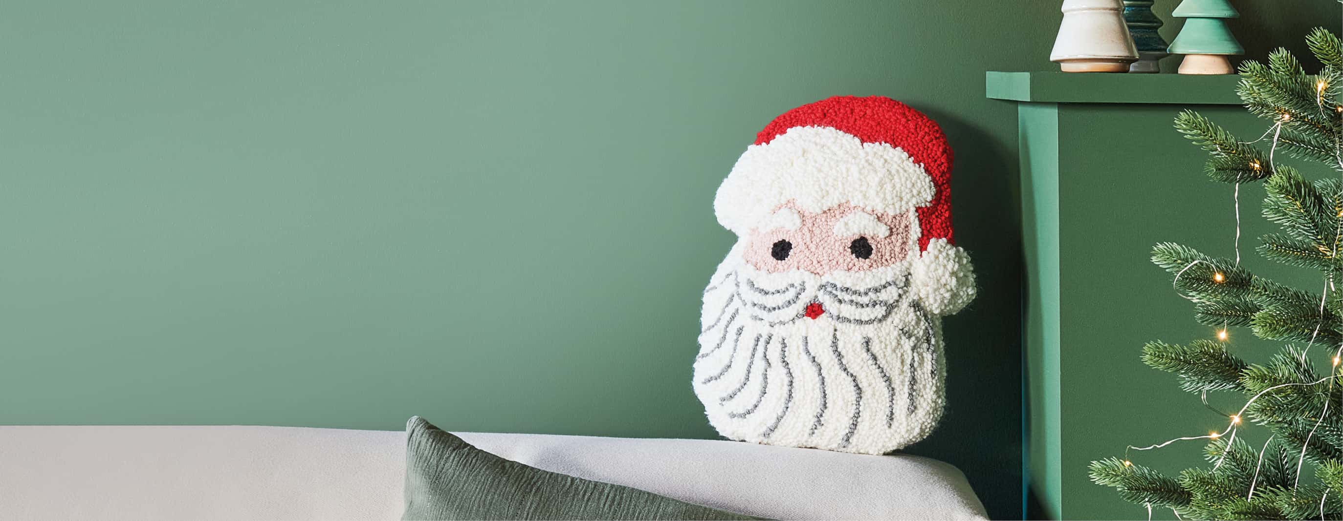 Punch-Needle-Kissen in Form eines Weihnachtsmanns auf einem Sofa vor grüner Wand neben einem festlich dekorierten Weihnachtsbaum.