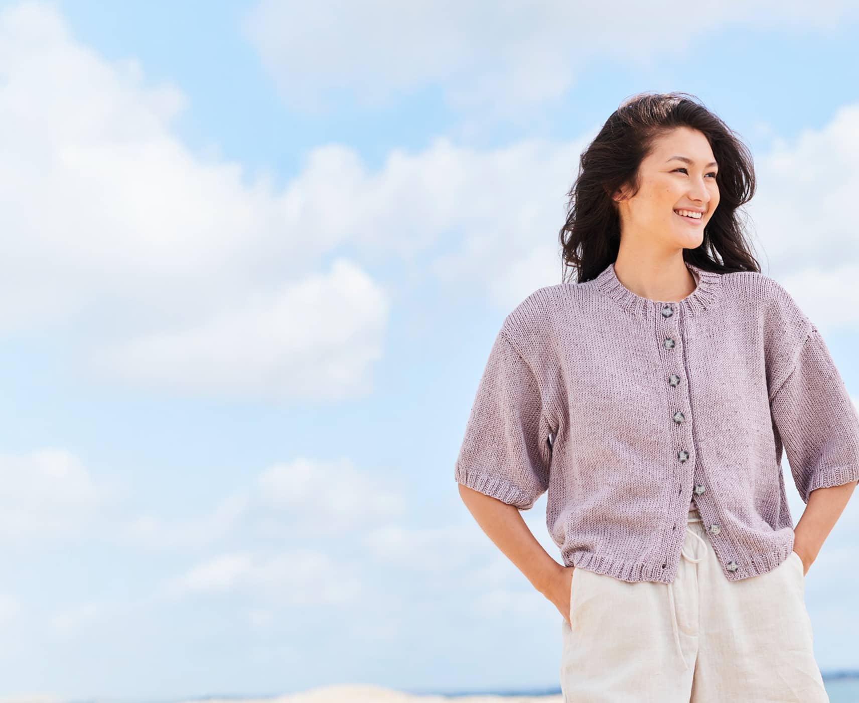 Eine Frau trägt eine gestrickte Kurzarmjacke. Im Hintergrund ist ein blauer Himmel mit Wolken.