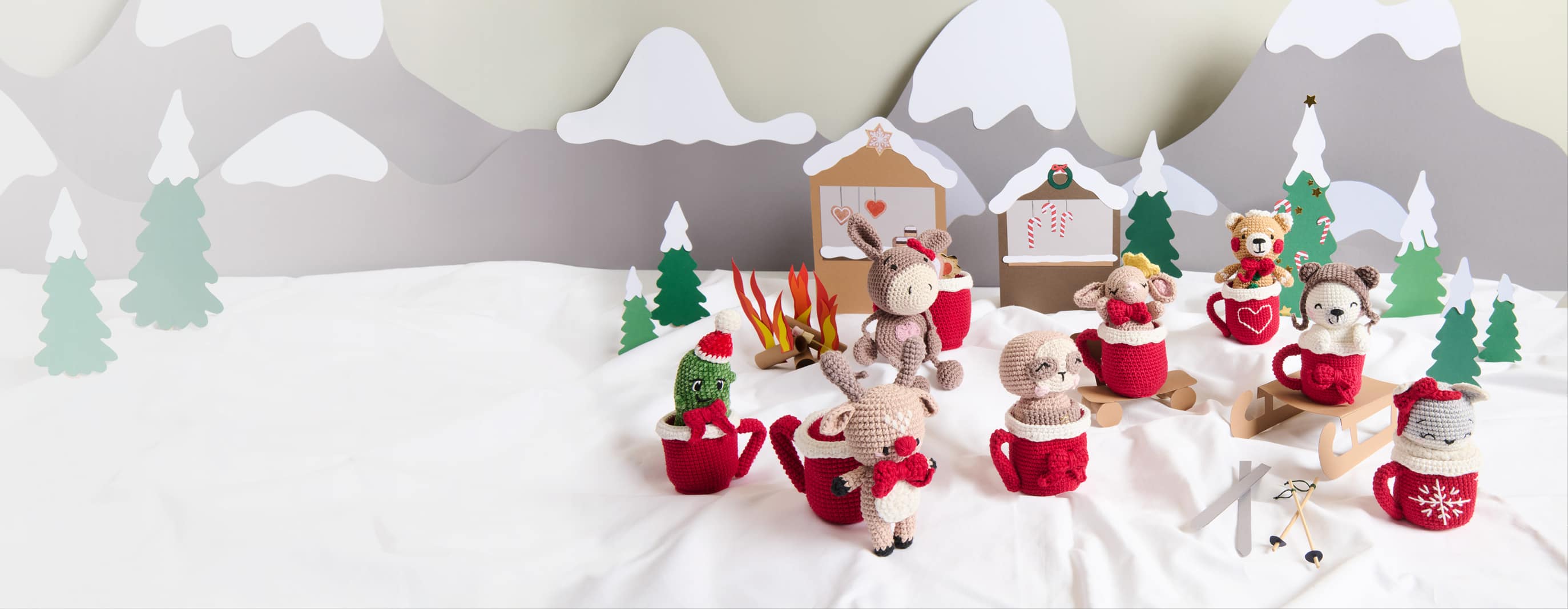 Weihnachlich geschmückte Amigurumis mit gehäkelten Tassen sind in einer aus Papier gestalteten Winterlandschaft dekoriert. 