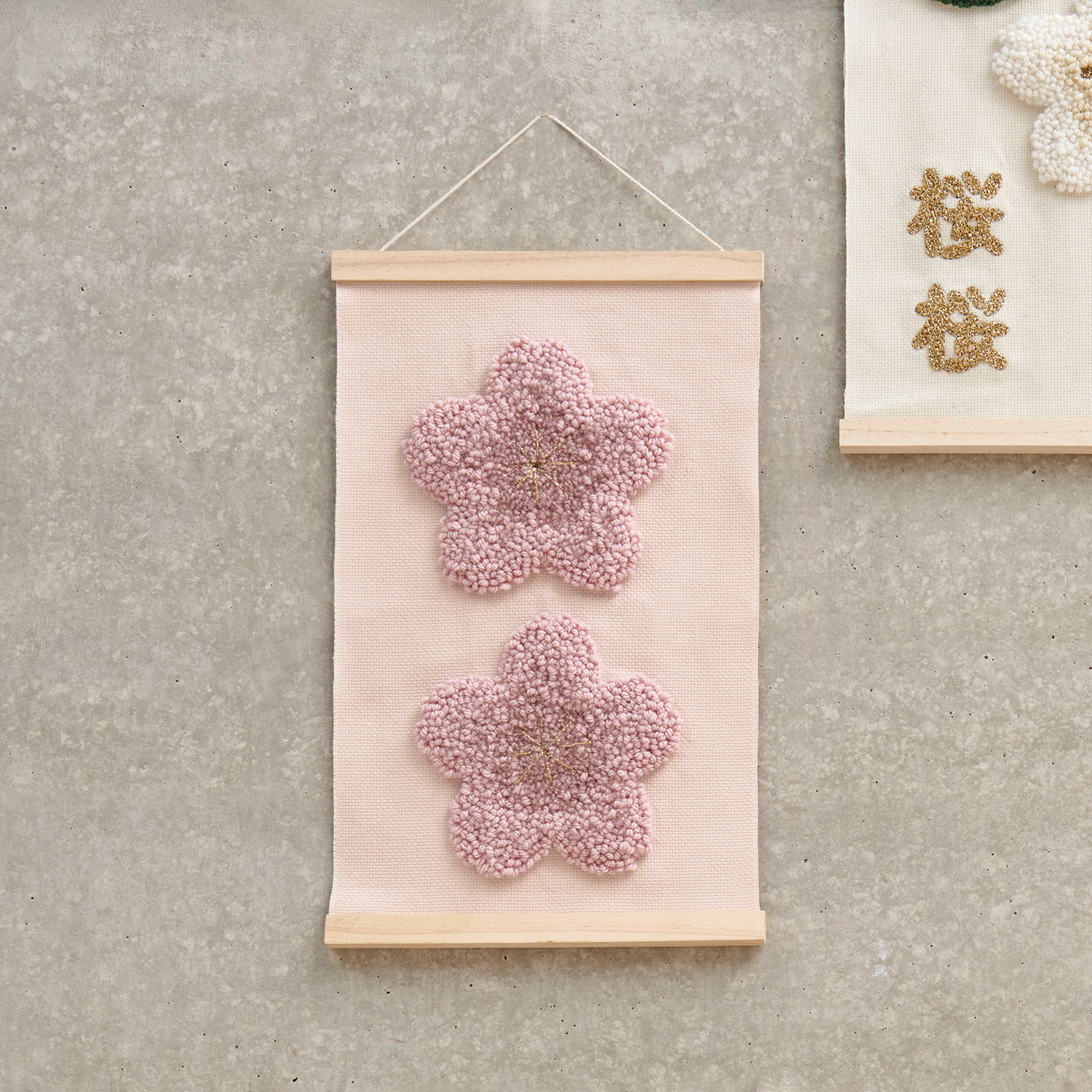 Anleitung Punch Needle Wandbehang mit rosa Blüten zeigt zwei dekorative Blumen auf beigem Stoff.