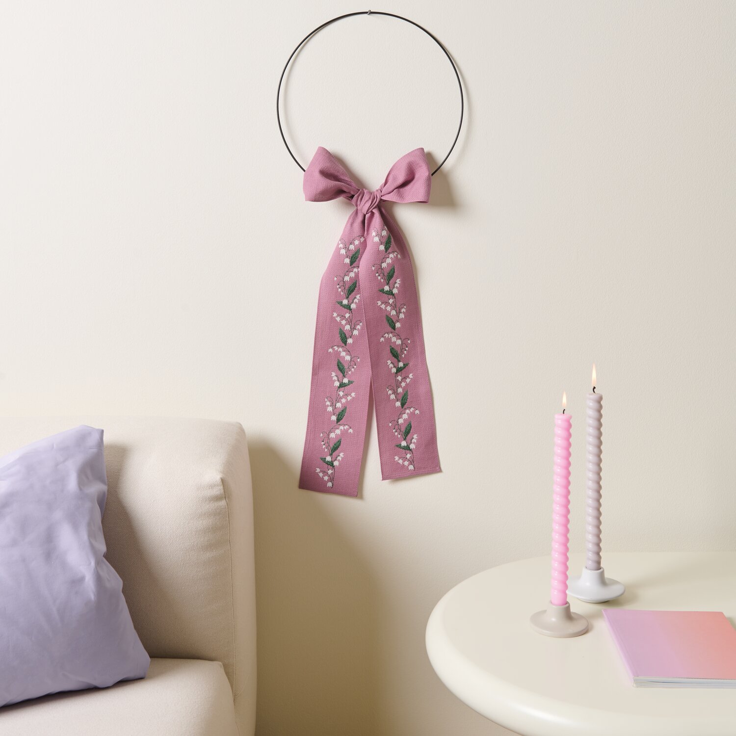 Stickpackung Gezählter Kreuzstich Schleife Maiglöckchen, rosa, dekoriert mit Maiglöckchen-Muster, hängt an einer Wand über einem Sofa.