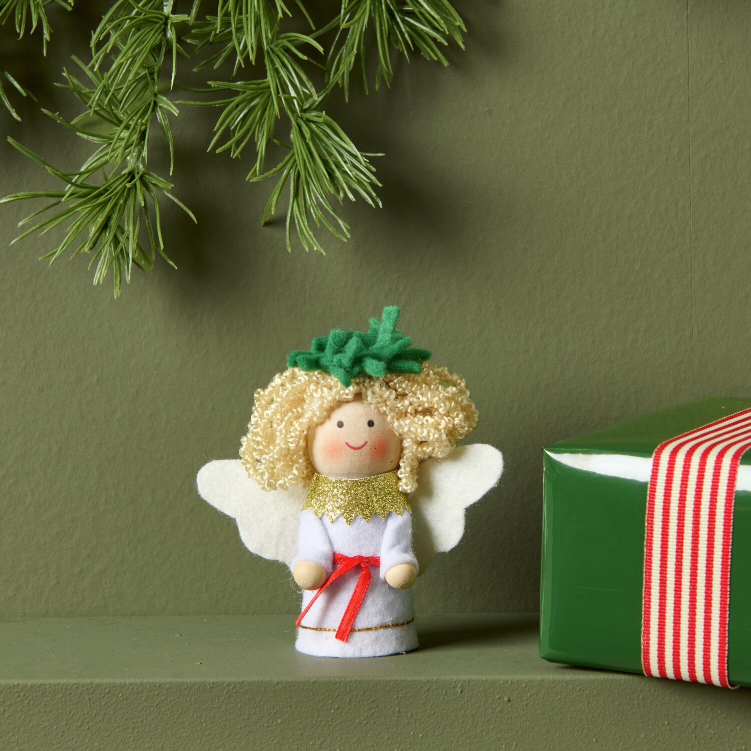Engel mit Filzkleid Weiß: Niedliche Figur mit goldenen Locken, grüner Krone und rotem Band, neben grünem Geschenk auf Regal.