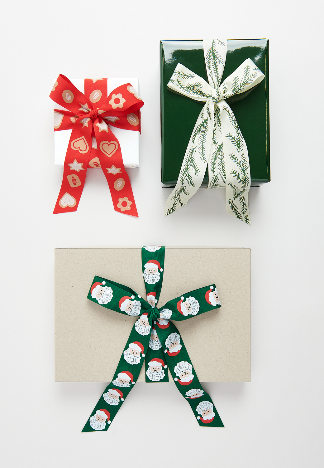 Drei Geschenke mit Schleifen verziert liegen auf grauem Hintergrund. Die Schleifenbänder zeigen weihnachtliche Designs mit Plätzchen, Zweigen und Weihnachtsmännern
