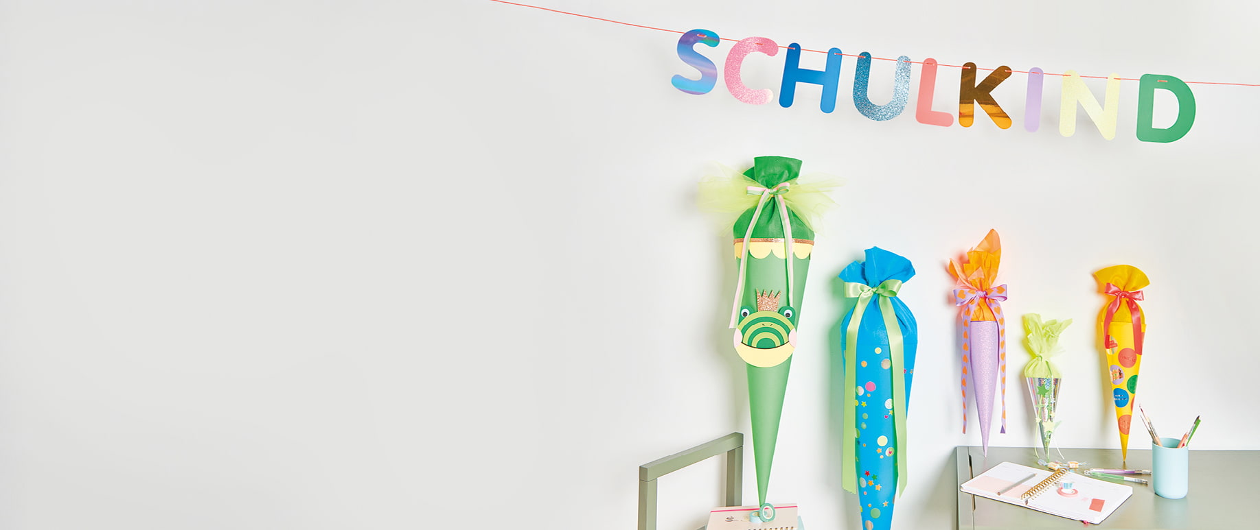 Gebastelte Schultüten in verschiedenen Größen und Farben stehen aufgestellt nebeneinander. Darüber eine Girlande mit dem Wort Schulkind