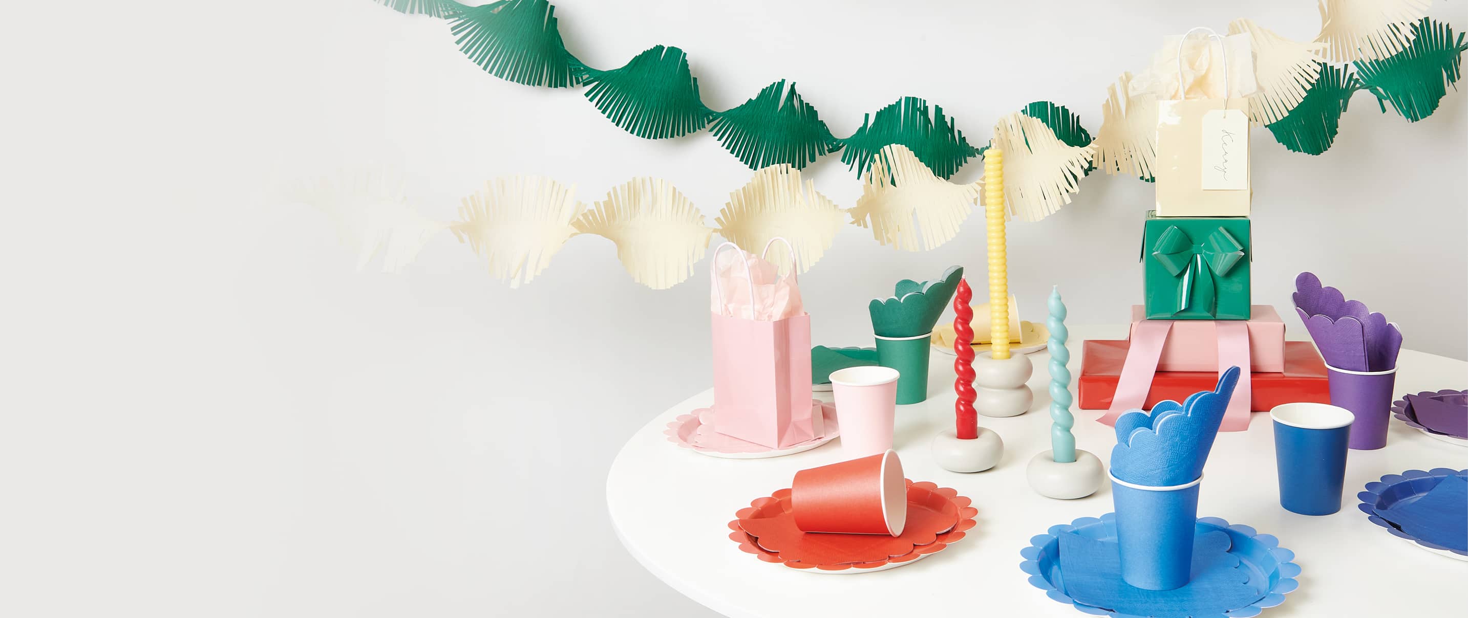 Gedrehte Kreppgirlanden hängen über einem gedeckten Tisch mit farblich abgestimmten Party-Sets aus Servietten, Tellern und Bechern in rot, grün, blau, lila