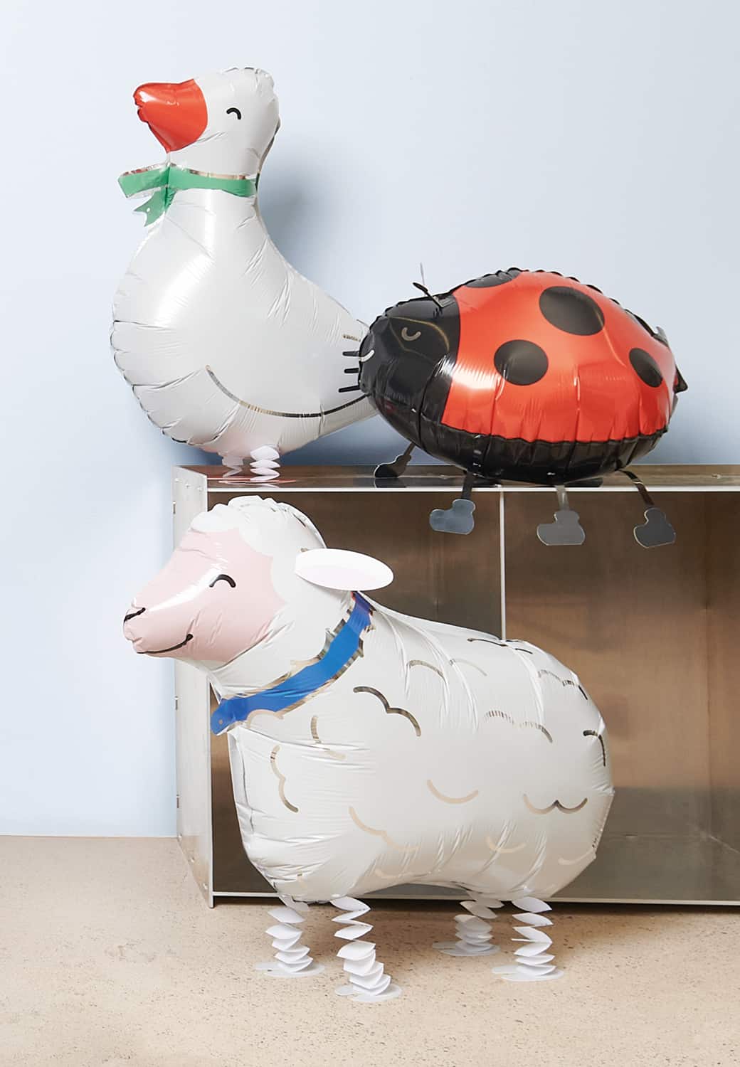 Drei Folienballons Airwalker in Form eines Schafs, einer Gans und eines Marienkäfers sind vor einem Regal aufgestellt.