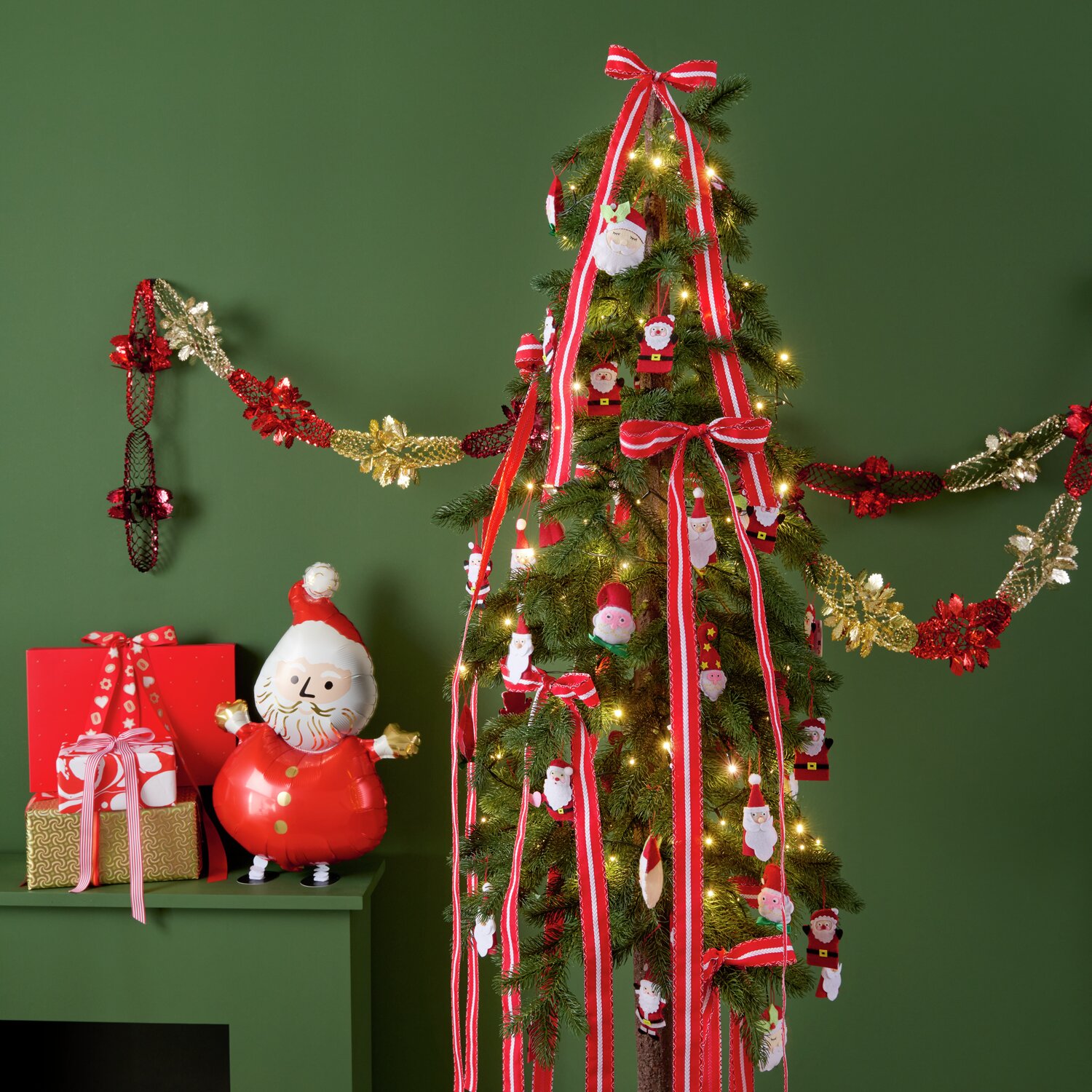 Auf dem Bild ist ein festlich geschmückter Weihnachtsbaum mit roten und weißen Filz-Santa-Aufhängern von Rico Design zu sehen. Der Baum ist mit Lichtern und Schleifen dekoriert. Links stehen Geschenke und eine Santa-Figur.