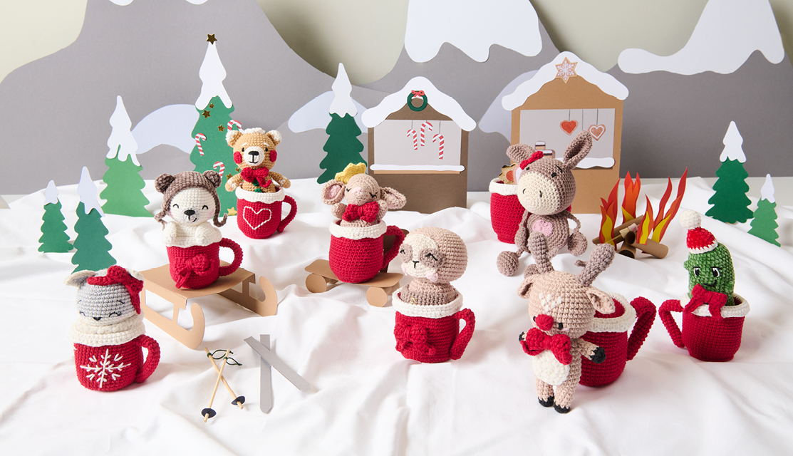 Häkelanleitung Ricorumi CAL Gary the Gingerbread Bear Häkelanleitung Ricorumi CAL Gary the Gingerbread Bear: Bunte Amigurumi-Tiere in roten Tassen vor winterlicher Kulisse mit Schnee und Tannen.