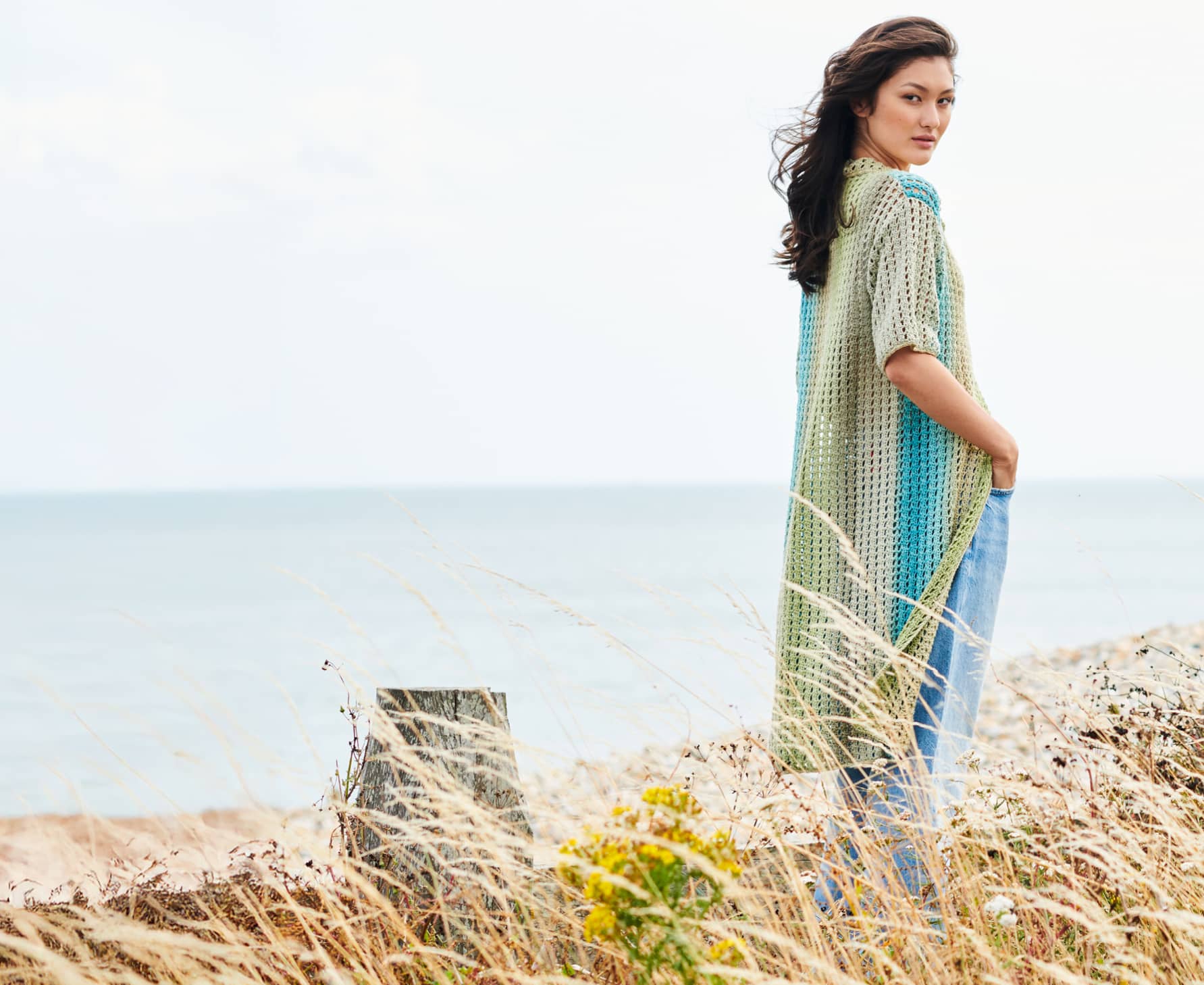 Eine Frau trägt eine gehäkelte, lange Jacke und steht in einer Düne. 