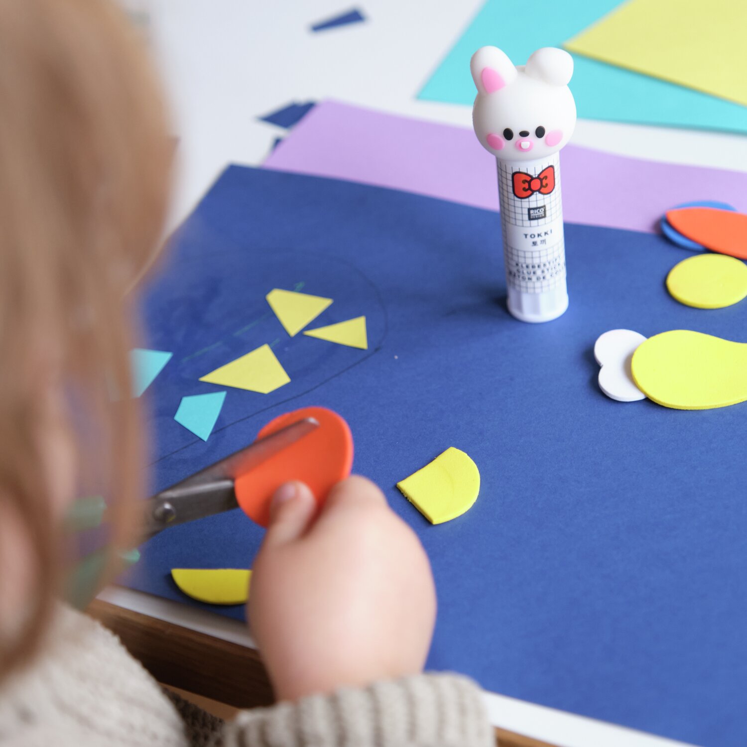Paper Poetry Klebestift Hase Weiß auf Basteltisch mit buntem Papier und Kind beim Schneiden.