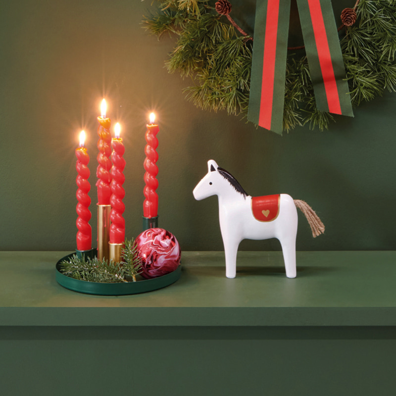 Ein Adventskranz mit roten Kerzen, ein Holzpferd und ein Kranz stehen dekoriert auf einem grünen Kaminsims 