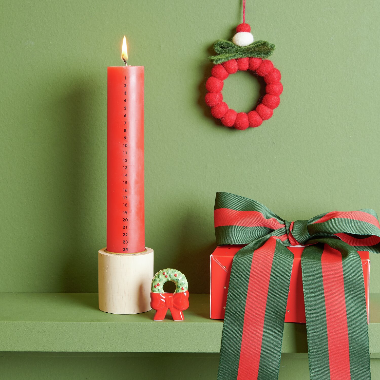 Porzellan Kranz mit Schleife Grün/Rot auf grünem Regal, neben roter Kerze und Geschenkbox.
