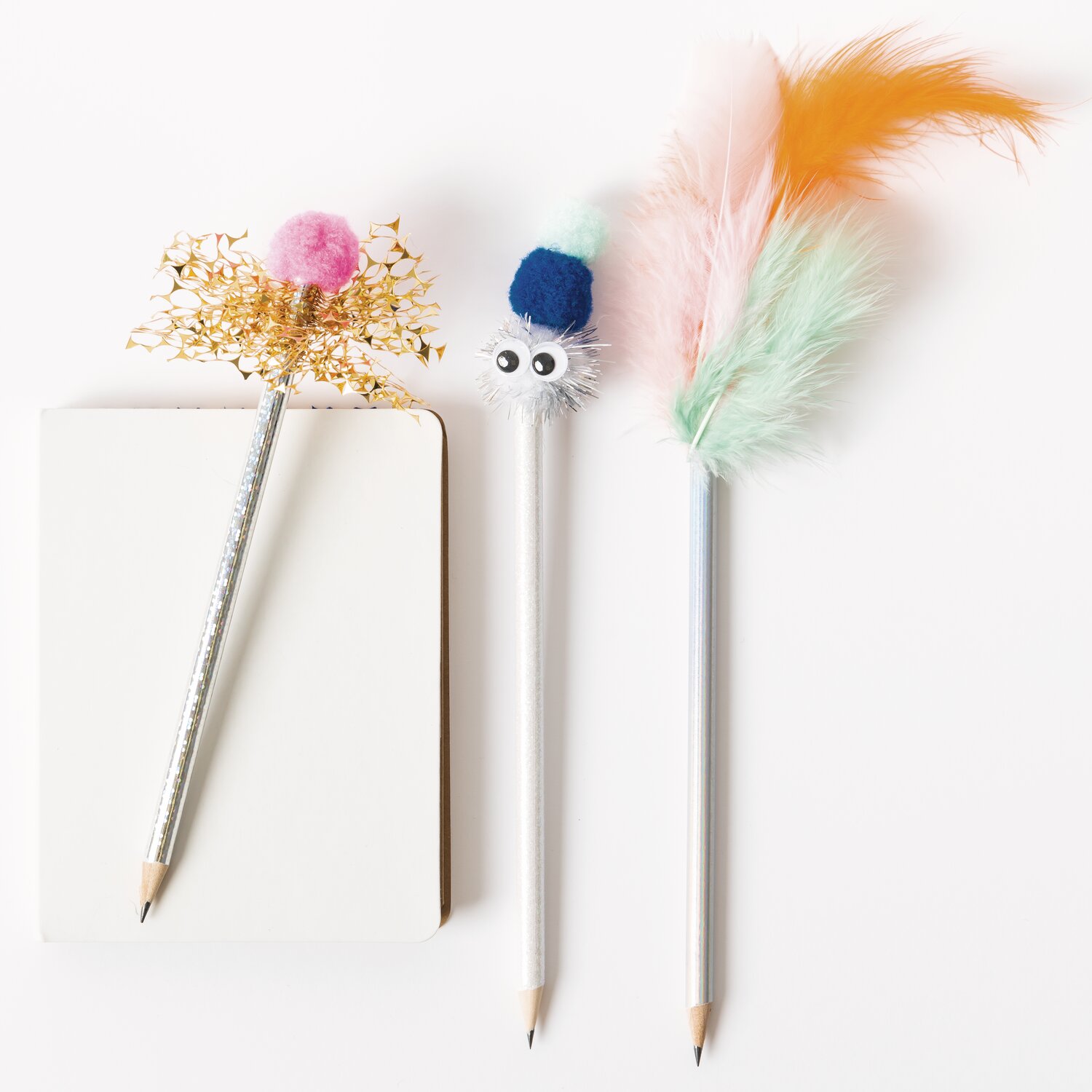 DIY-Bastelset bunt: Drei verzierte Stifte mit Federn und Pompons neben einem Notizblock.