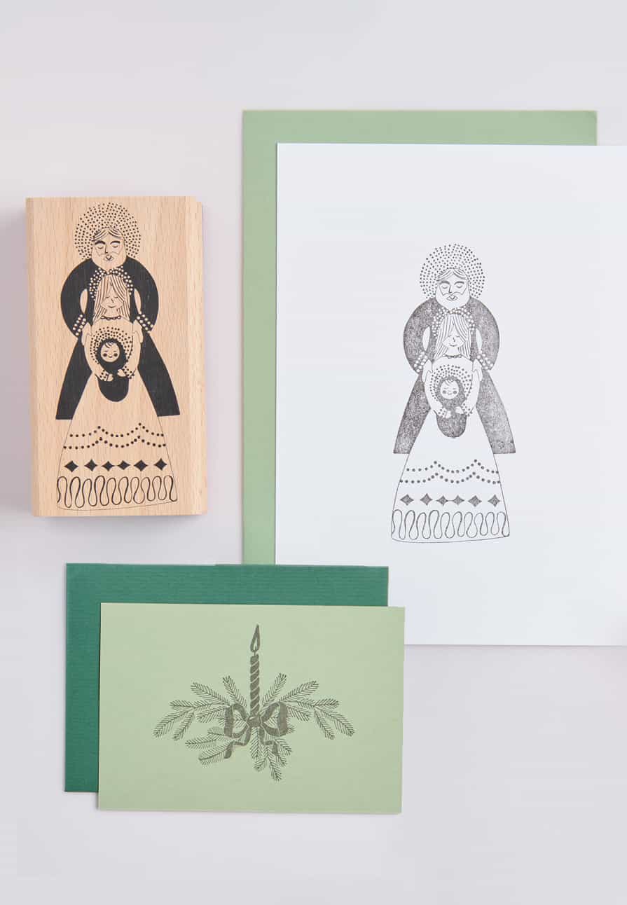 Ein Holzstempel mit Motiv der Holy Family liegt neben bestempelten Karten auf hellgrauem Hintergrund. 