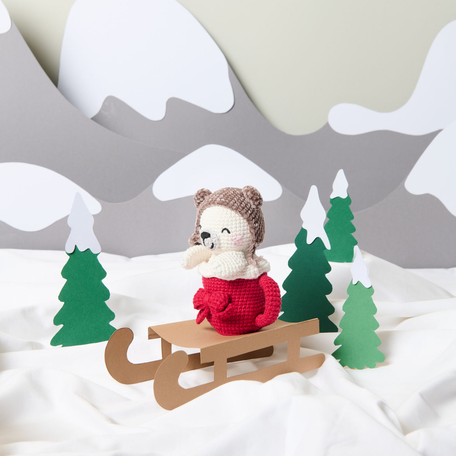 Häkelanleitung Ricorumi CAL Sam the Christmas Seal: Niedliche gehäkelte Robbe auf einem Schlitten, umgeben von grünen Tannen und schneebedeckten Bergen.