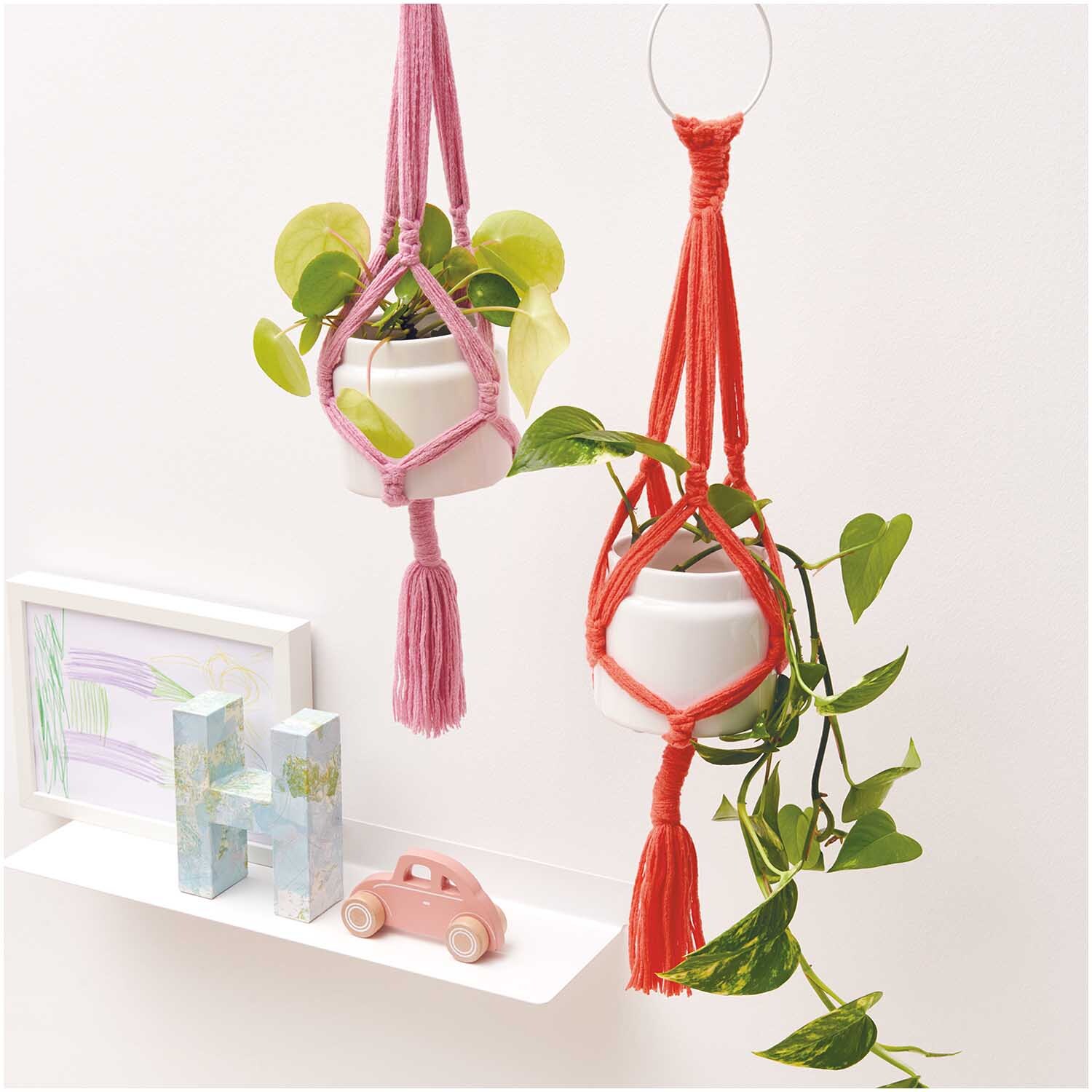 Creative Loopy: Zwei Makramee-Blumenampeln in Rosa und Rot mit Pflanzen, dekorativ im Raum hängend.