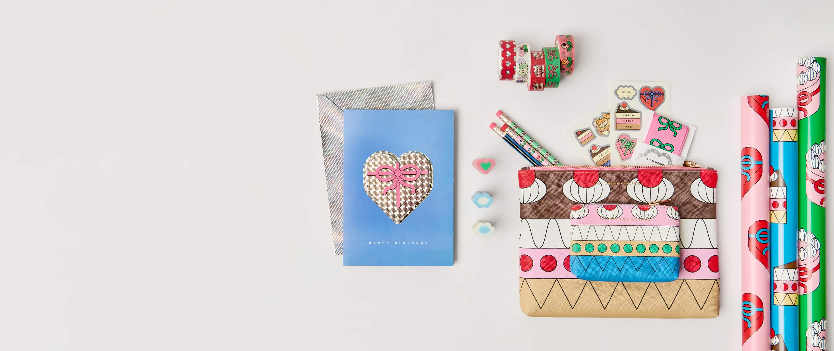 Bunte Karte, Etuis, Geschenkpapier und Schreibwaren-Artikel mit Illustrationen von Gebäck, Herzen und Schleifen aus der Kollektion Eye Candy liegen auf grauem Hintergrund.