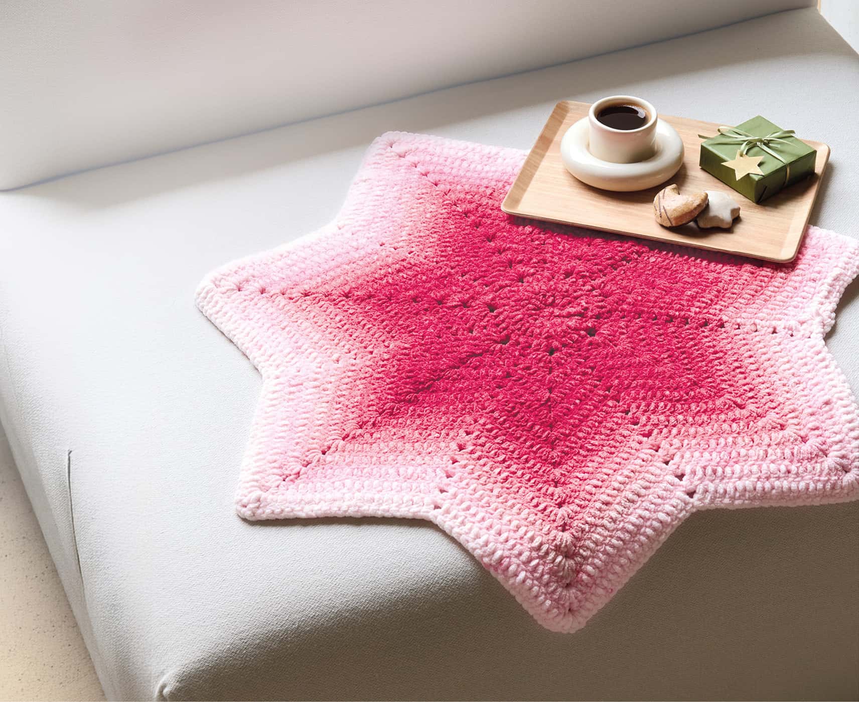 Eine gehäkelte, sternförmige Decke mit rotem Farbverlauf liegt auf einem Sofa.
