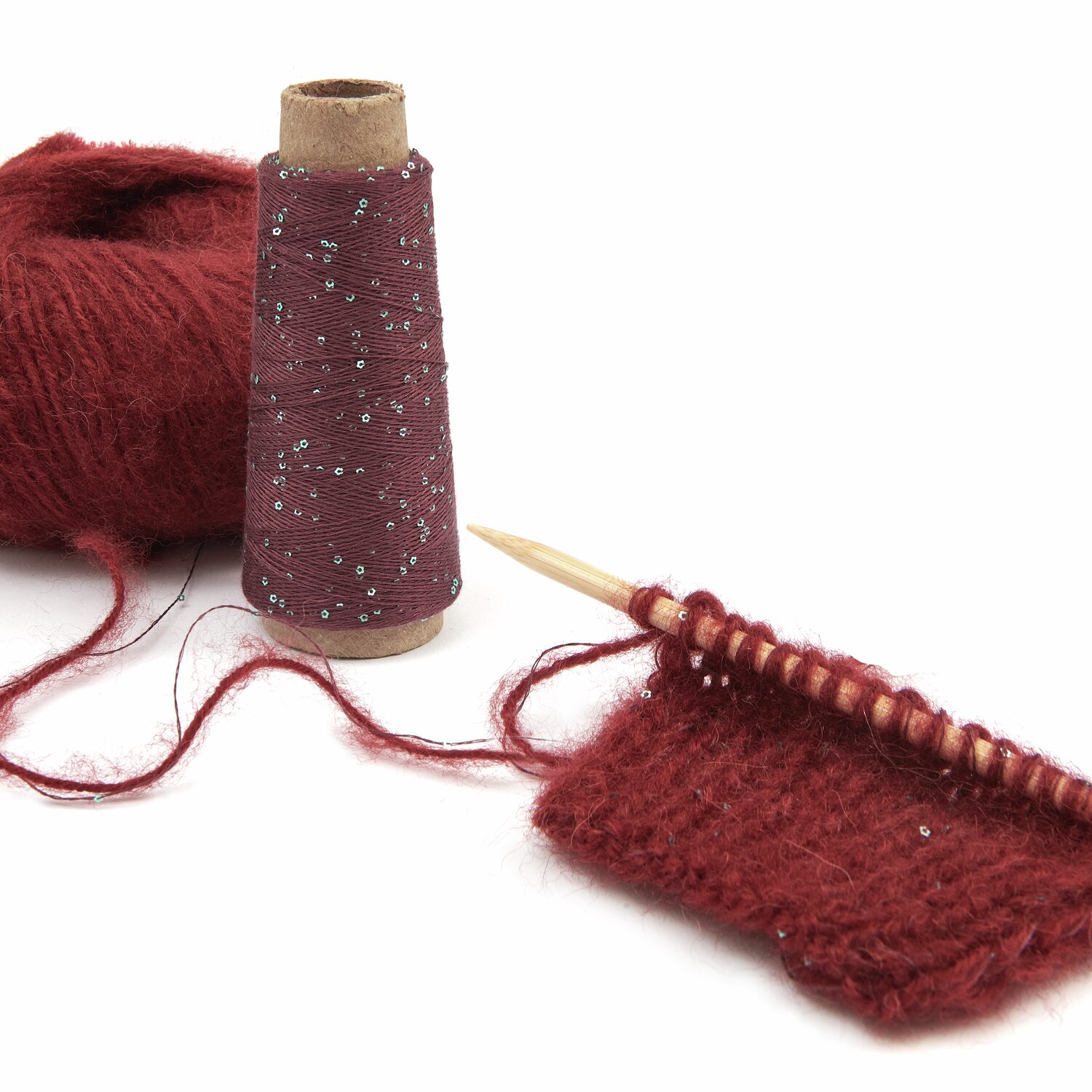 Rotes Strickgarn mit Glitzerfaden, auf einer Holzstricknadel gestrickt. Daneben eine Garnrolle. Perfekt für kreative Strickprojekte. Marke: Unbekannt.