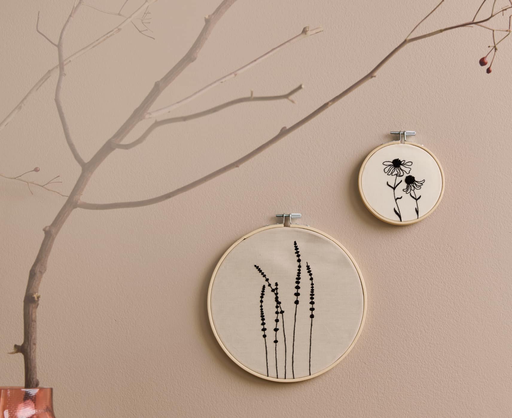 Stickbilder mit Gräser- und Blumenmotiven hängen an einer beigen Wand. 