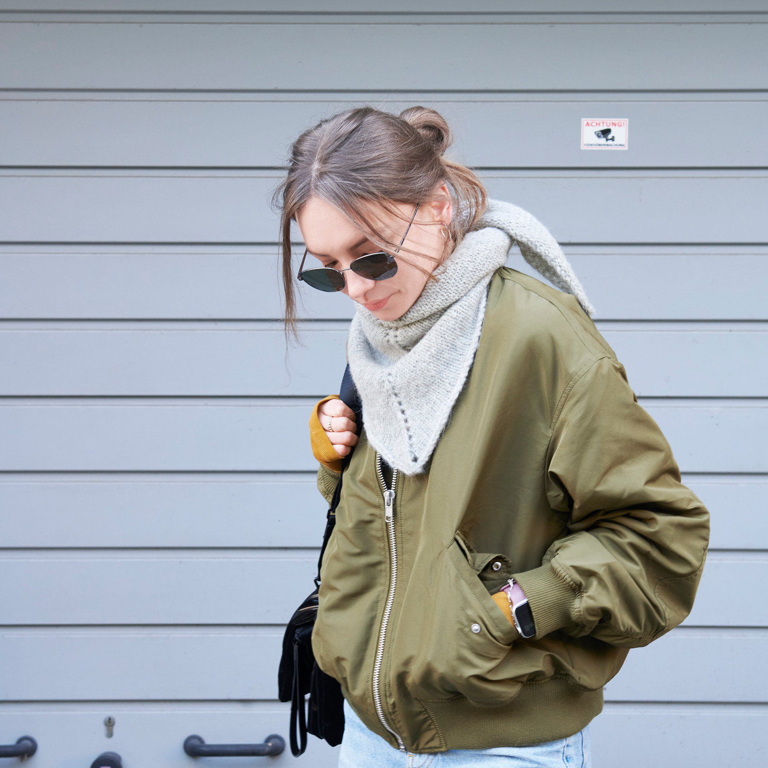 Strickset Dreieckstuch Modell 05a: Frau mit olivgrüner Jacke und grauem Tuch vor grauer Wand.