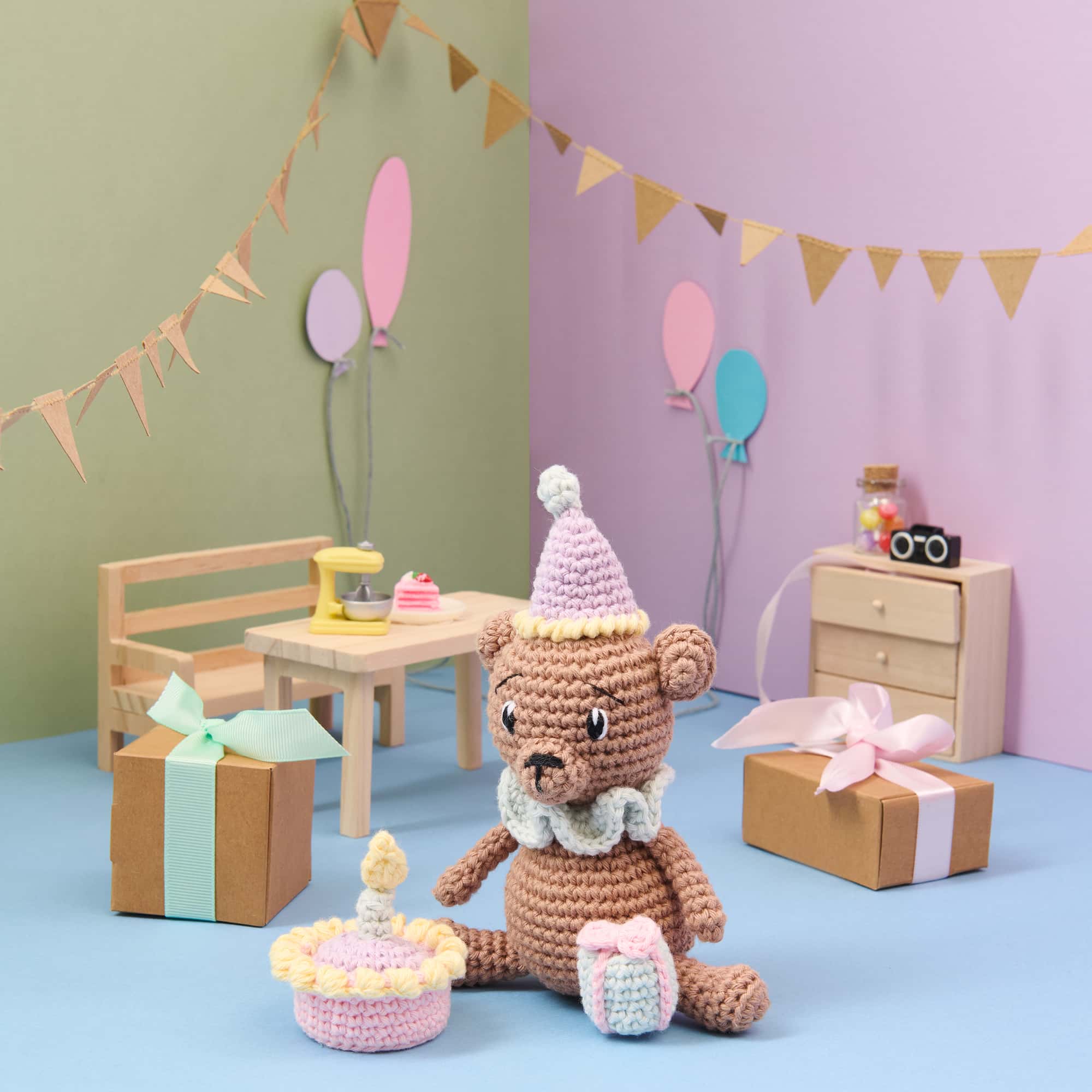 Ein gehäkelter Bär mit Partyhut und Geschenk sitzt vor einer gehäkelten Torte. Um ihn herum ist eine bunt dekorierte Szene mit Partydekoration aufgebaut.