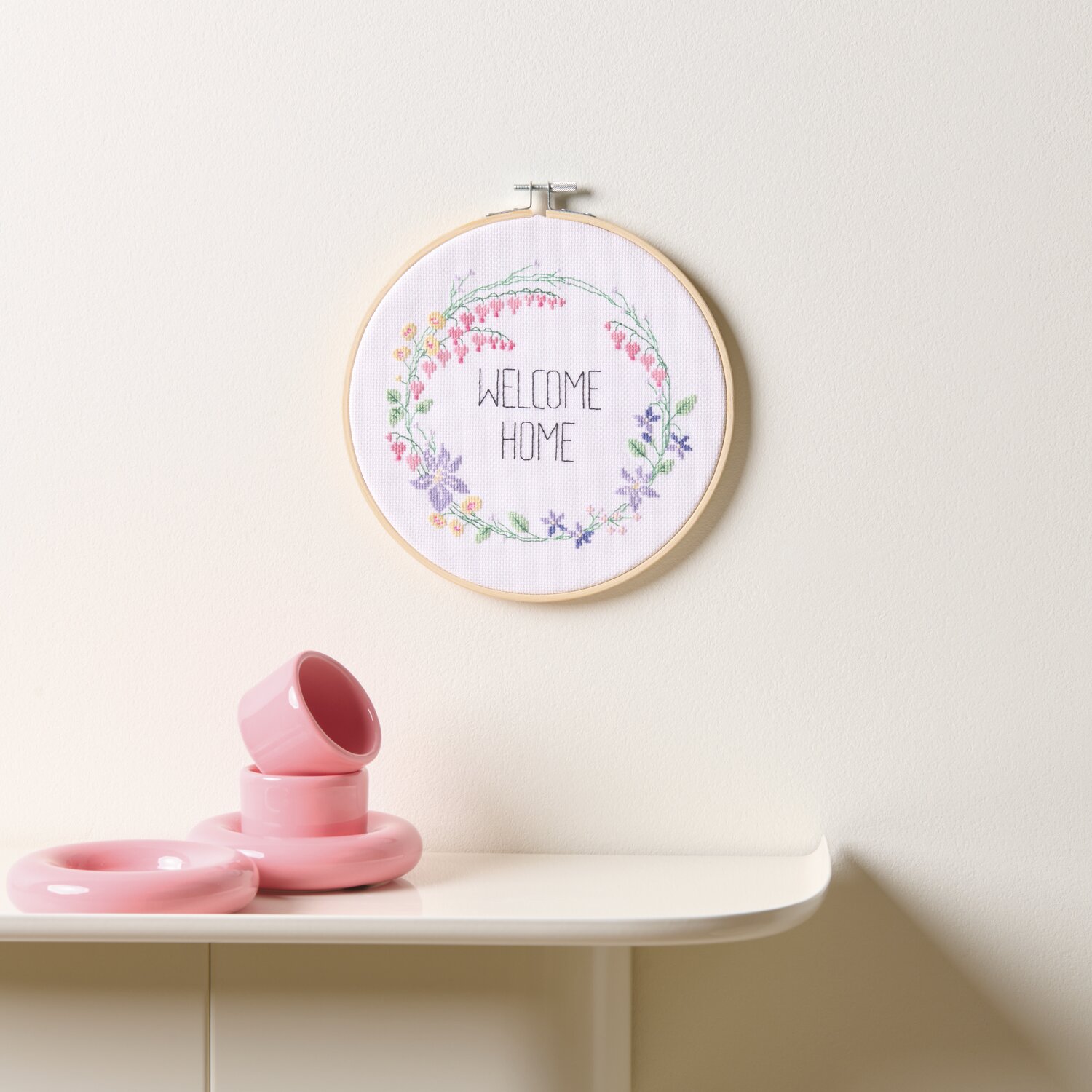 Stickpackung Gezählter Kreuzstich Blumenkranz, Ø20cm, zeigt florales Design an weißer Wand. Darunter rosa Keramik auf Tisch.