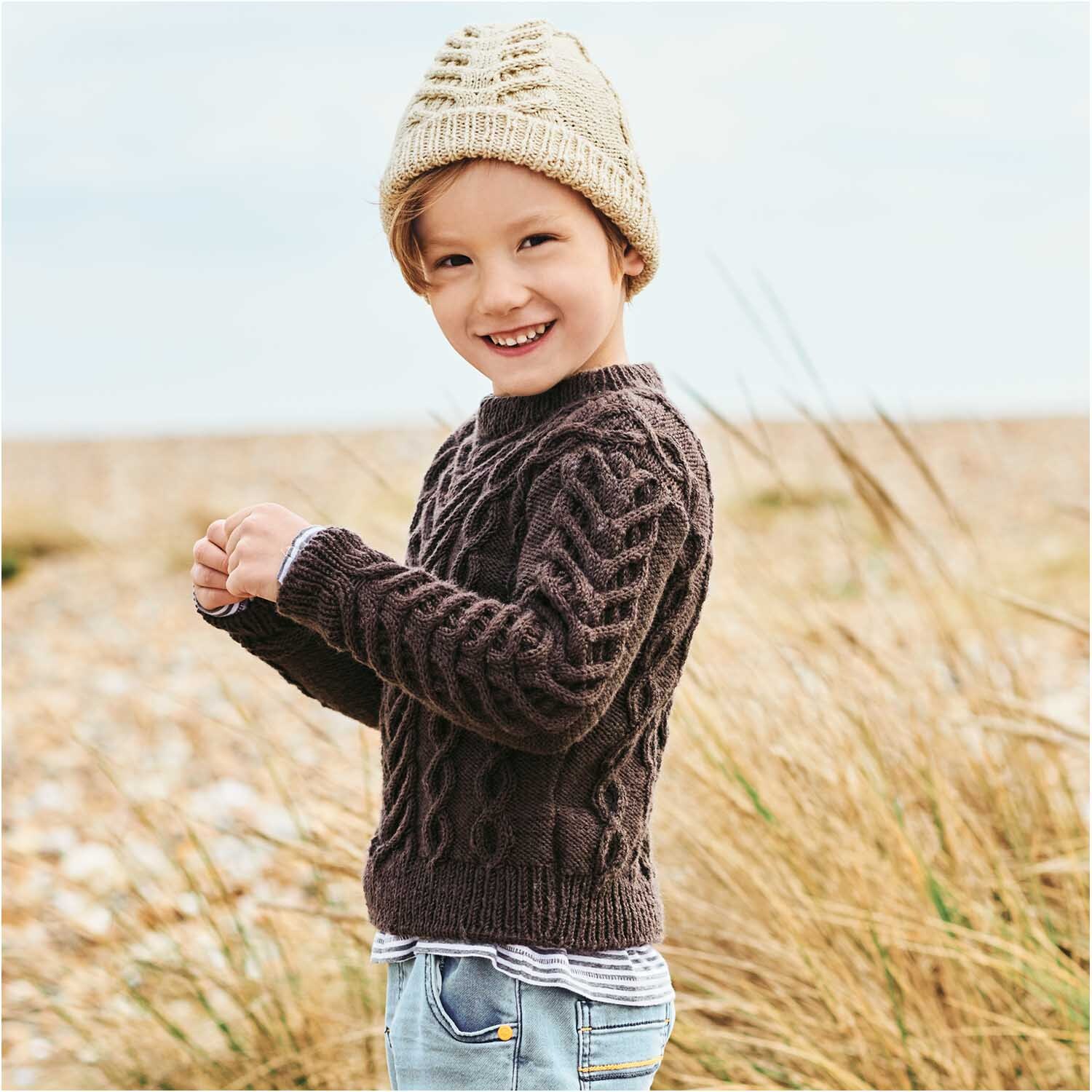 Essentials Merino dk: Kind in gestricktem Pullover am Strand, trägt eine Mütze, Rico Design.