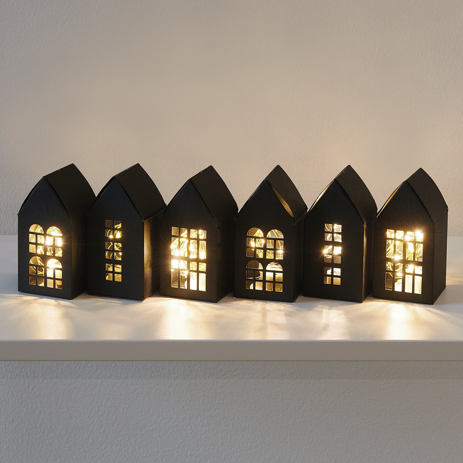 Paper Poetry Papphäuser-Set: Sechs schwarze, beleuchtete Papphäuser auf einem Regal, ideal als Adventskalender.