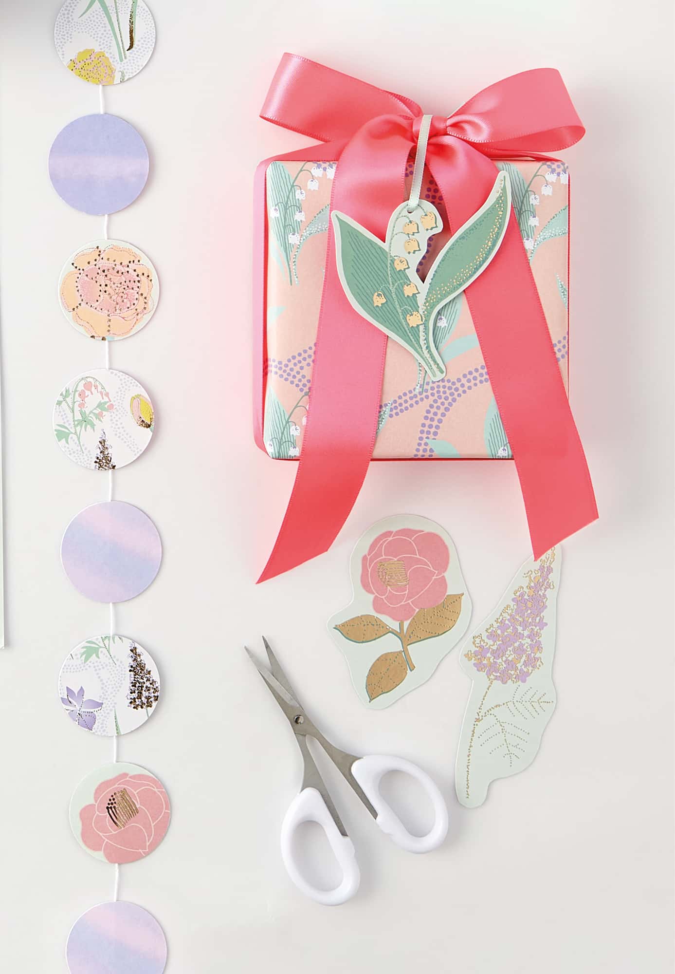 Eine Papiergirlande aus Kreisen, ein eingepacktes Geschenk mit floralen Geschenkanhängern und eine Schere liegen auf hellem Hintergrund. 