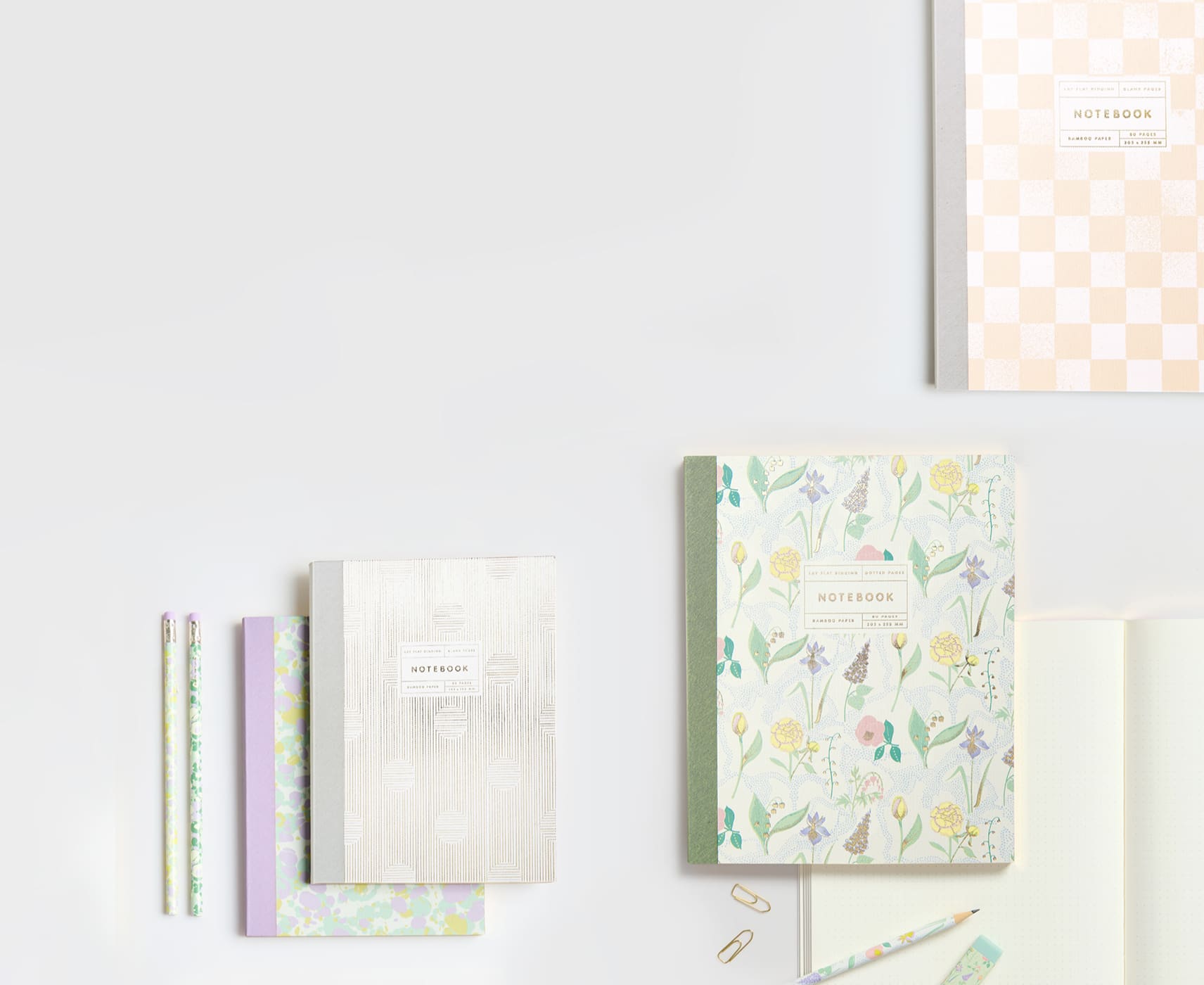 Notizbücher in Pastelltönen, floralen und abstrakten Mustern liegen umgeben von Bleistiften auf grauem Hintergrund.