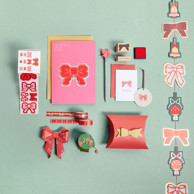 Papeterie-Produkte wie Karten, Sticker und Stempel mit Schleifen-Designs liegen arrangiert auf grünem Hintergrund