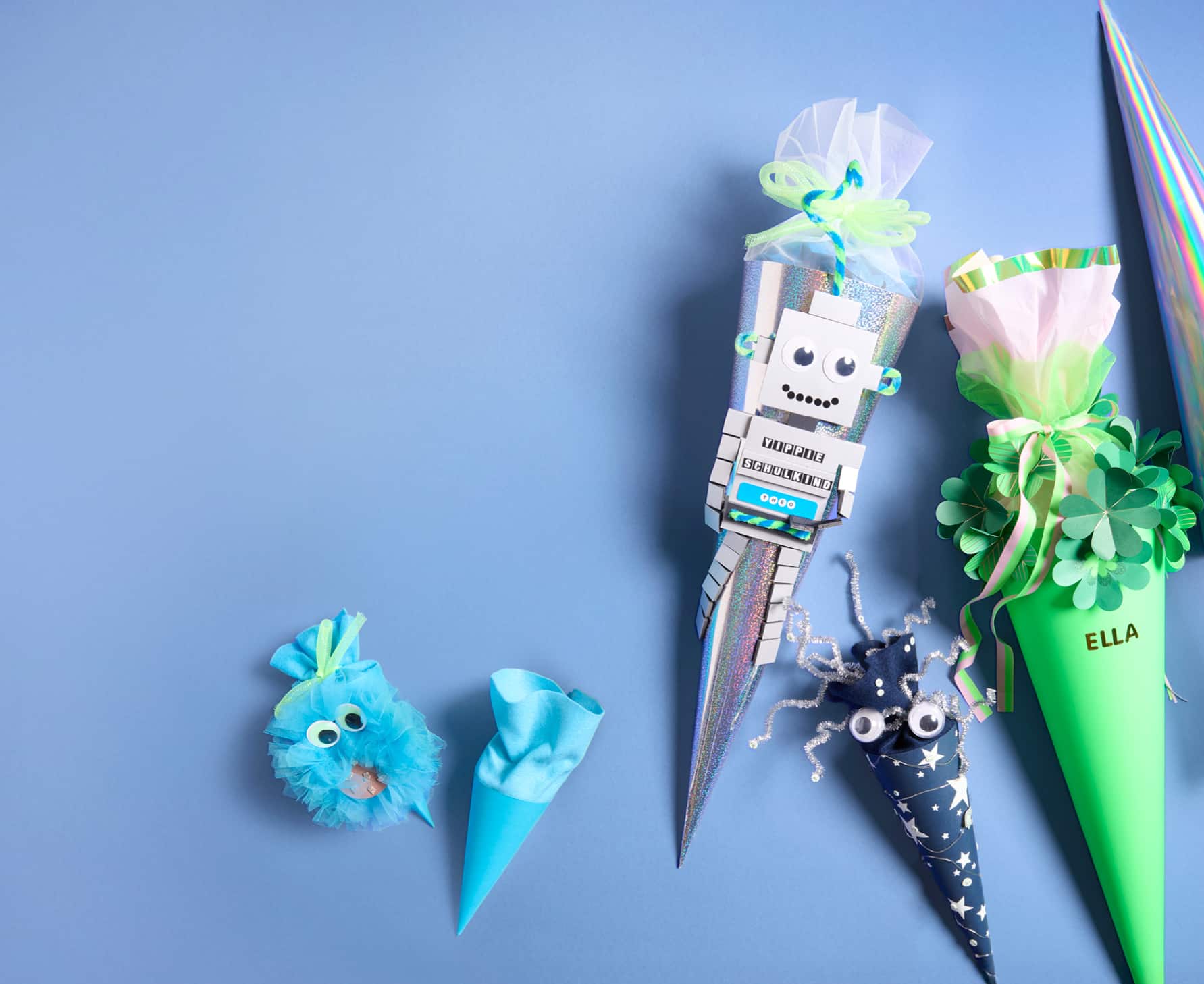 Gebastelte Schultüten in verschiedenen Größen und Designs liegen auf blauem Hintergrund. Eine Schultüte ist mit Roboter-Motiv, eine weitere ist grün mit Kleeblättern 