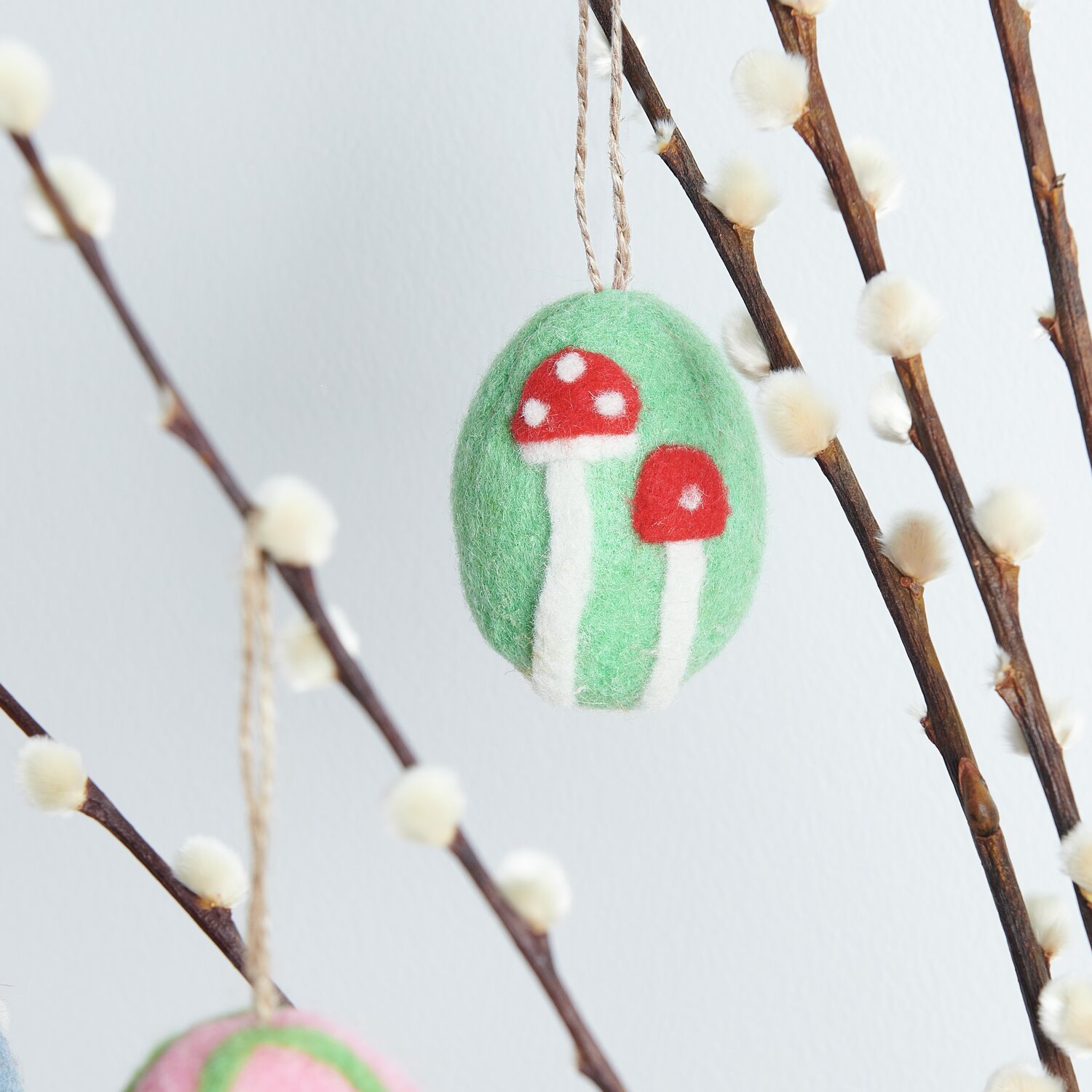 Aufhänger Filz-Ei Purpur-Blume: Grünes Filz-Ei mit roten Pilzen, dekorativ an einem Zweig hängend.