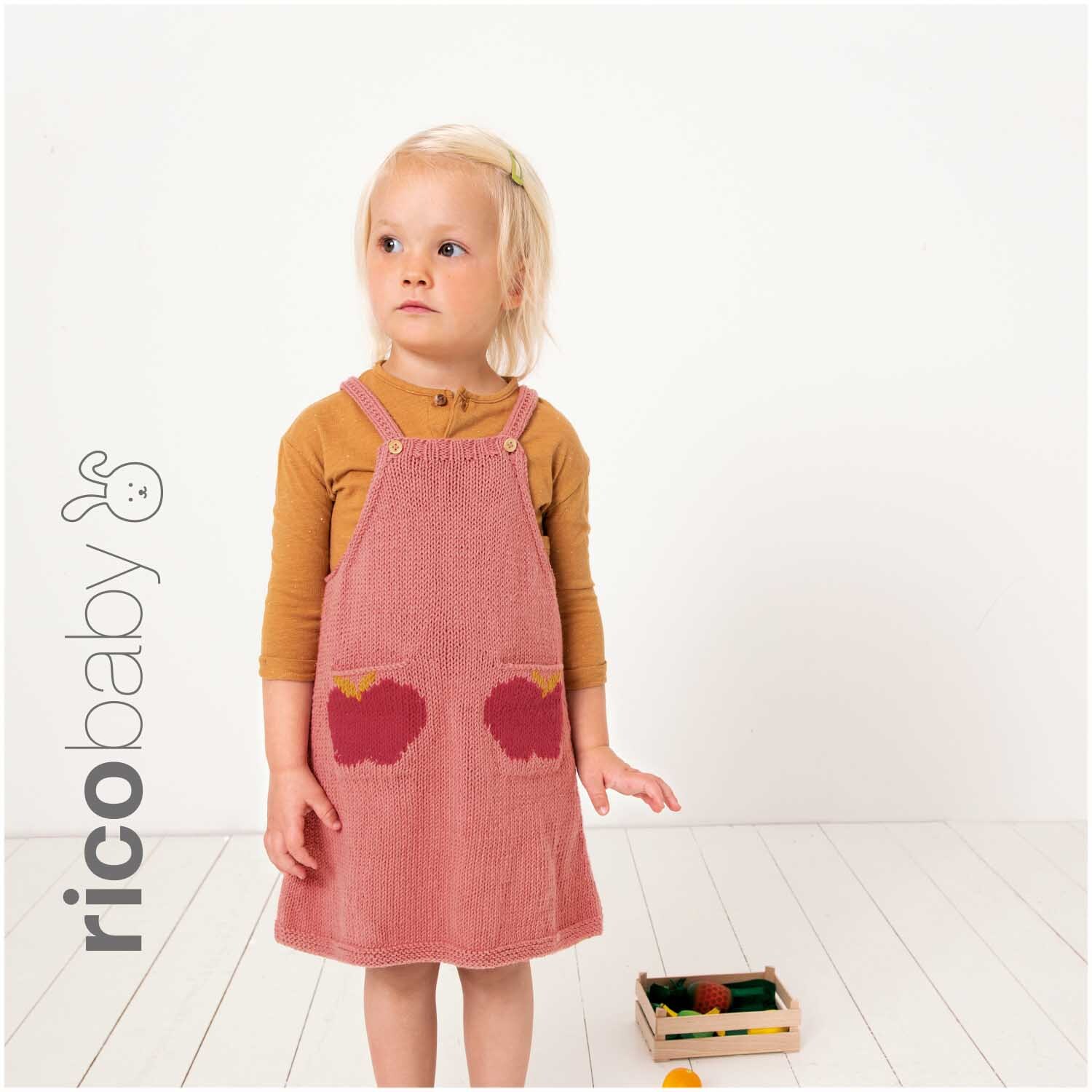 Rico Baby Cotton Soft dk: Kind in rosa Latzkleid mit Apfelmotiv, stehend auf hellem Boden.
