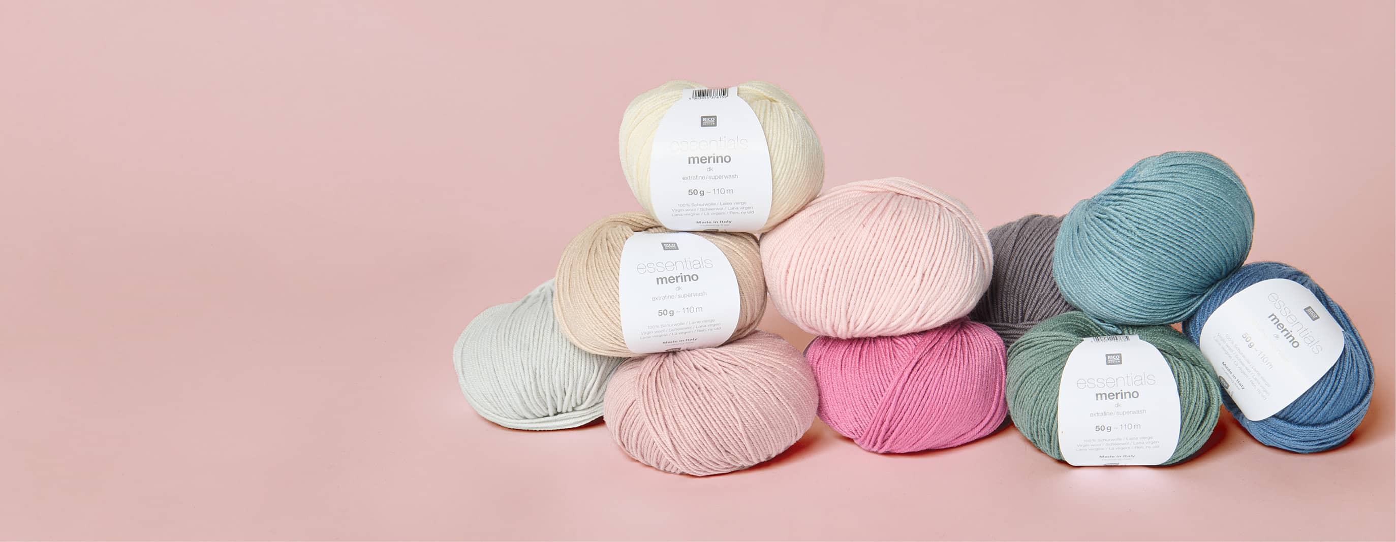 Mehrere Knäuel von Essentials Merino dk in verschiedenen Farben liegen gestapelt vor rosa Hintergrund. 